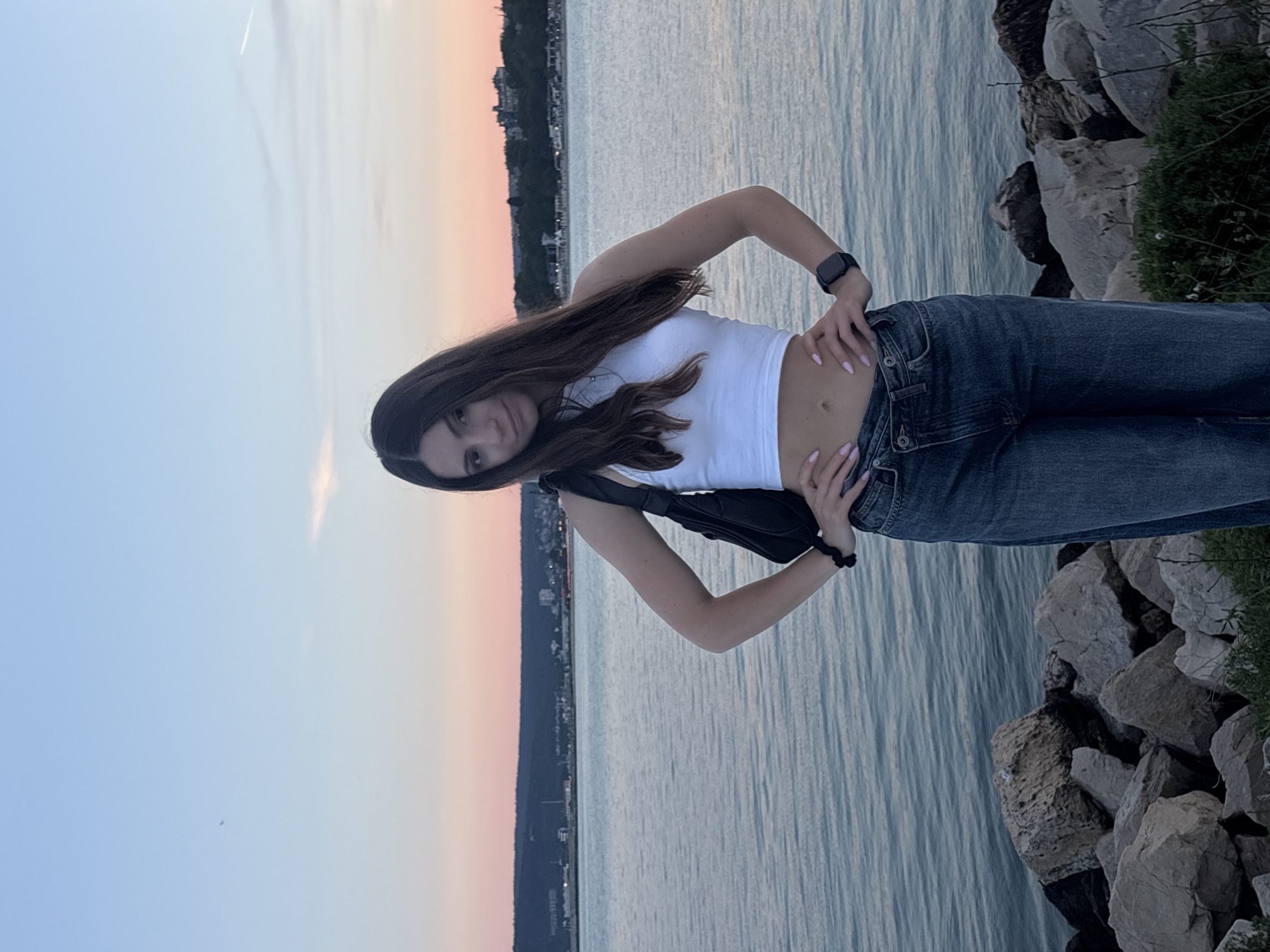 A young woman with long, dark hair stands on a rocky shoreline with a body of water behind her. She is wearing a white crop top, blue jeans, and a black shoulder bag. The sky is a gradient of blue and pink, suggesting a sunset. In the background, a city skyline is visible across the water, which appears to be Varna, Bulgaria. The woman is posing with her hands on her hips, looking at the camera.