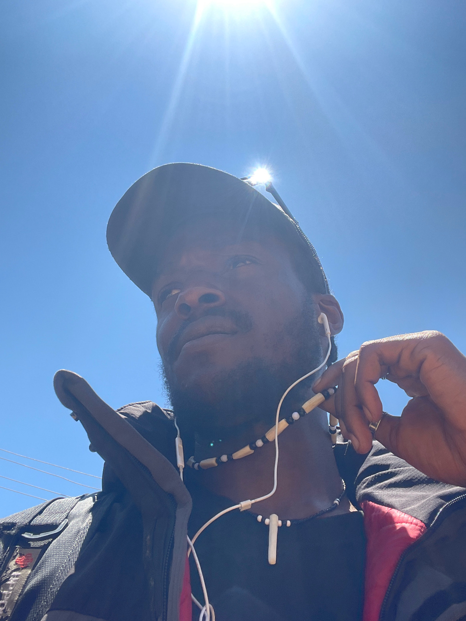 A low-angle, close-up shot captures a Black man wearing a dark baseball cap, a black t-shirt, and a black jacket with red lining. He has a beard and is wearing white earbuds connected to a necklace with wooden and black beads and a white cross pendant. He is looking upwards, and his right hand is touching his ear. The background is a bright blue sky with the sun directly overhead, creating lens flare. The image appears to have been taken outdoors, possibly on a sunny day in Jos, Nigeria, given the clear sky and bright sunlight typical of the region. There are power lines visible in the upper left background.