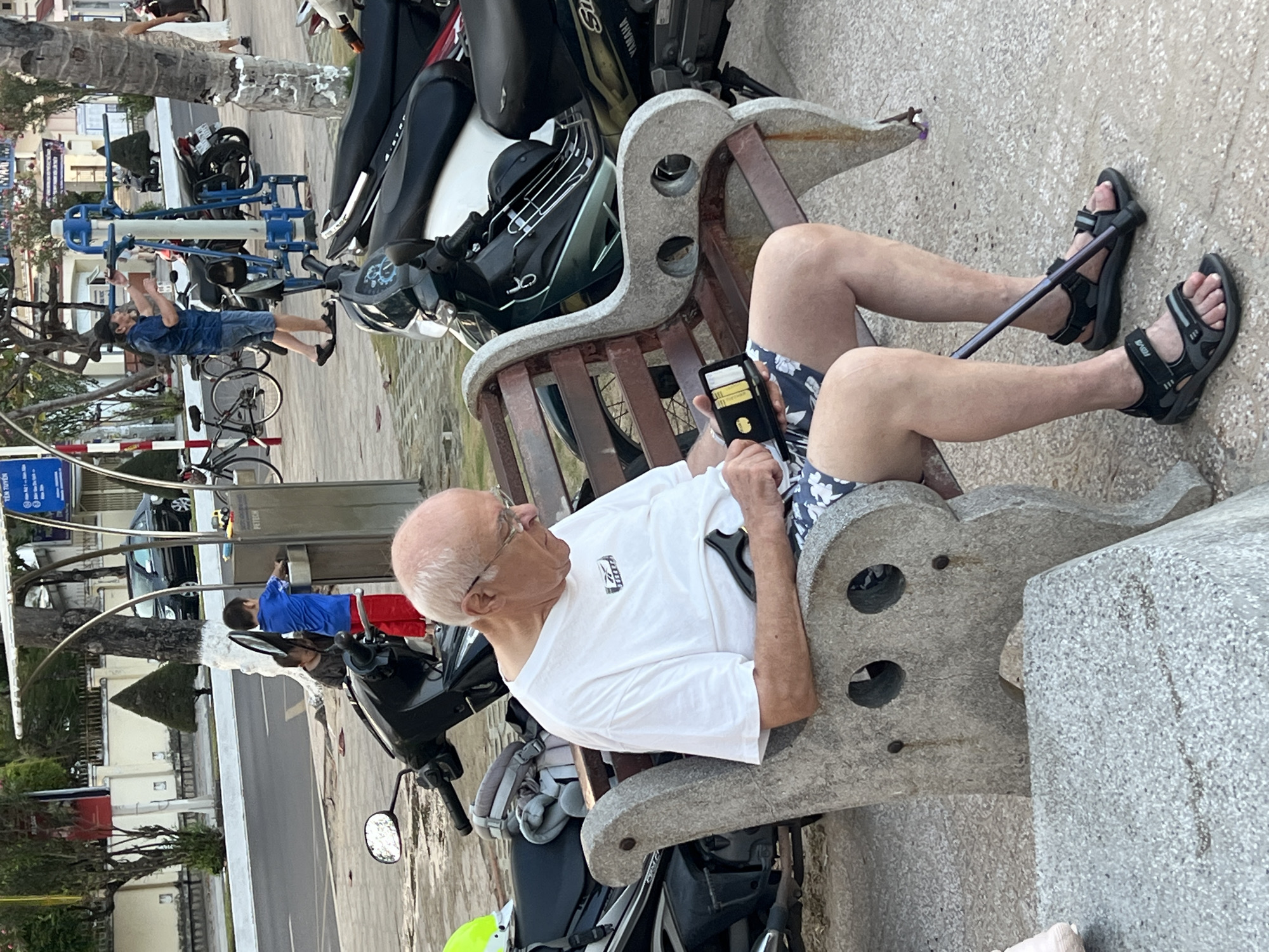 An outdoor scene in Bắc Nha Trang, Vietnam, captured in a vertically oriented, 90-degree clockwise rotated photograph. In the foreground, an older, bald man with glasses, wearing a white t-shirt, patterned shorts, and "NOVA" brand sandals, sits on a stone bench with wooden slats. He holds a black wallet or case containing small items, possibly cigarettes, and a walking stick rests between his legs.
Behind him, several motorcycles and scooters are parked, one with "YAMAHA" visible on its seat. In the mid-ground, a younger man in a blue t-shirt and denim shorts uses blue outdoor exercise equipment. A bicycle is parked nearby. Further in the background, a person in a blue shirt and red shorts stands near a car wash or water station. Various trees, greenery, and buildings line the street. The lighting suggests it is daytime. Visible text includes Vietnamese words on street signs, such as "KHU VỰC C...", "TIÊN TÊN B... ĐƯỜNG ĐỊA P...", and "PETECH" in the distance.