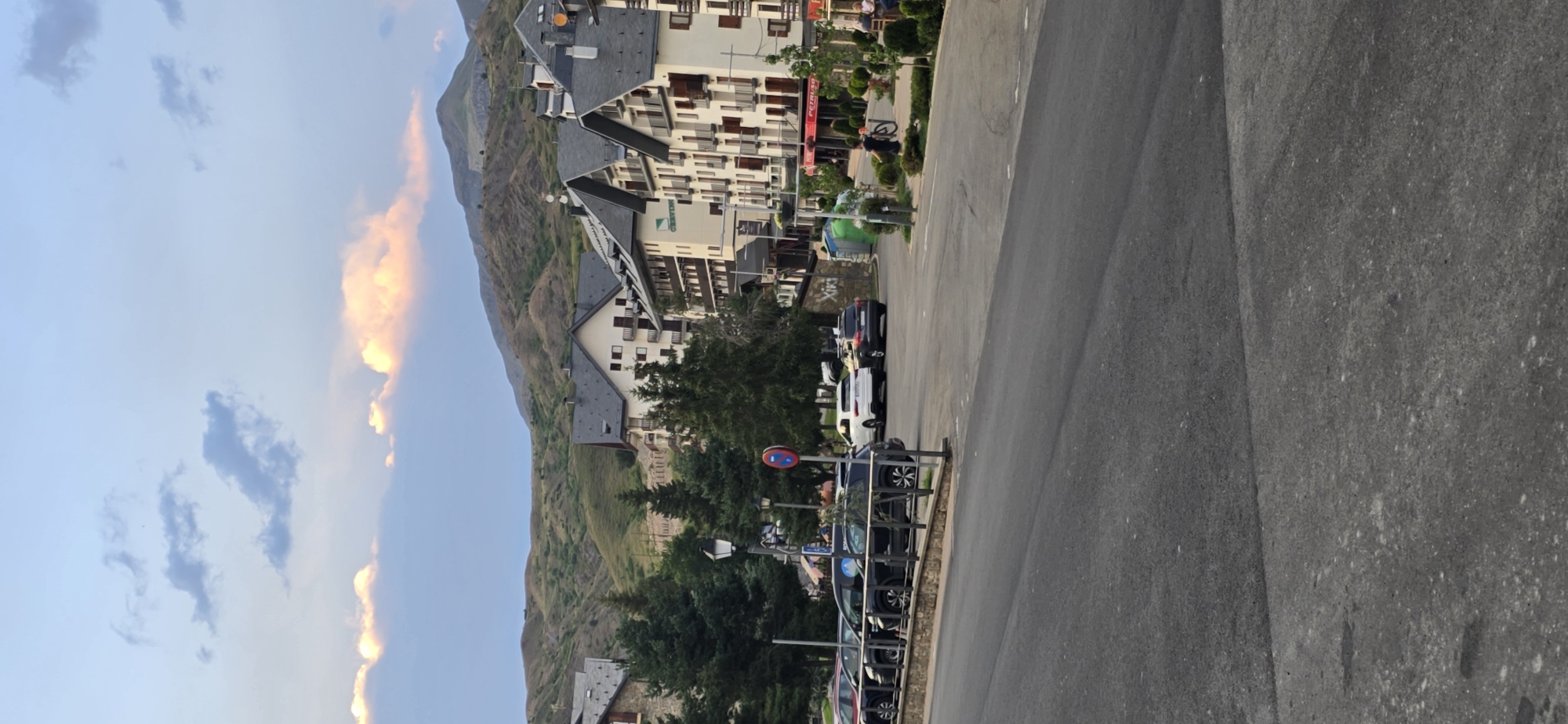 The image captures a view of Sallent de Gállego, Spain, a town nestled in the Pyrenees mountains. In the foreground, a paved road curves into the scene. To the left, parked cars line a street bordered by a low stone wall and landscaping. Beyond the cars and wall, several multi-story buildings with dark, sloped roofs and balconies are visible. Evergreen trees are interspersed between these buildings and in the mid-ground. In the background, rolling, green mountains rise under a sky with scattered clouds tinged with pink and orange, suggesting either sunrise or sunset. Several people are walking on the sidewalks in front of the buildings. One person is pushing a bicycle. The overall impression is a tranquil, mountain town setting.