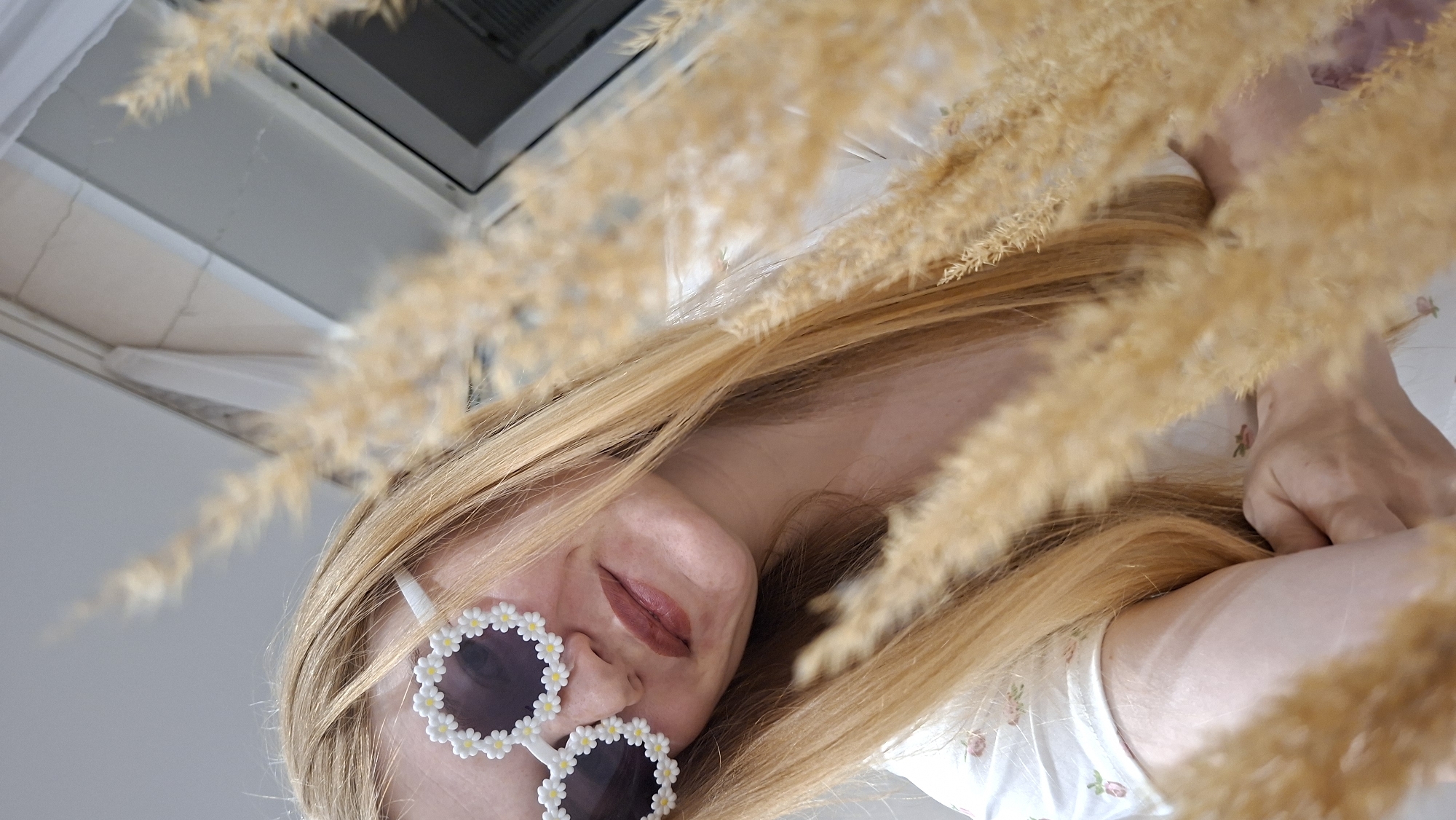 A vertically oriented, close-up photograph depicts a young adult female subject indoors, stated to be located in Chaikovsky, Russia. The subject is positioned in the left and central areas of the frame, with her head slightly tilted. She has long, straight, blonde hair that frames her face and rests on her shoulders. Her eyes are obscured by a pair of white, novelty sunglasses designed with daisy-shaped frames, featuring dark, circular lenses. The daisy petals are composed of small white beads, and the centers are yellow beads. Her mouth is slightly open in a subtle smile, with lips colored in a reddish-brown hue. She is wearing a light-colored, possibly white or cream, short-sleeved top adorned with a small, repeating pattern of pink or red floral motifs with green leaves.
The immediate foreground, particularly on the right side and bottom, is heavily blurred by a dense cluster of dried, feathery, light-brown/tan botanical elements, obscuring parts of the subject's right arm and torso. Her right hand is partially visible near her chest, though its exact action is indistinct due to the foreground elements.
The background is out of focus, revealing a light-colored wall or ceiling in the upper left quadrant. To the right, a white window or door frame is visible, with a darker, indistinct area beyond it, suggesting an exterior or a reflective surface. The lighting is bright and diffuse, appearing to originate from the background window area. No specific urban or landmark identifiers for Chaikovsky are visible within the image.