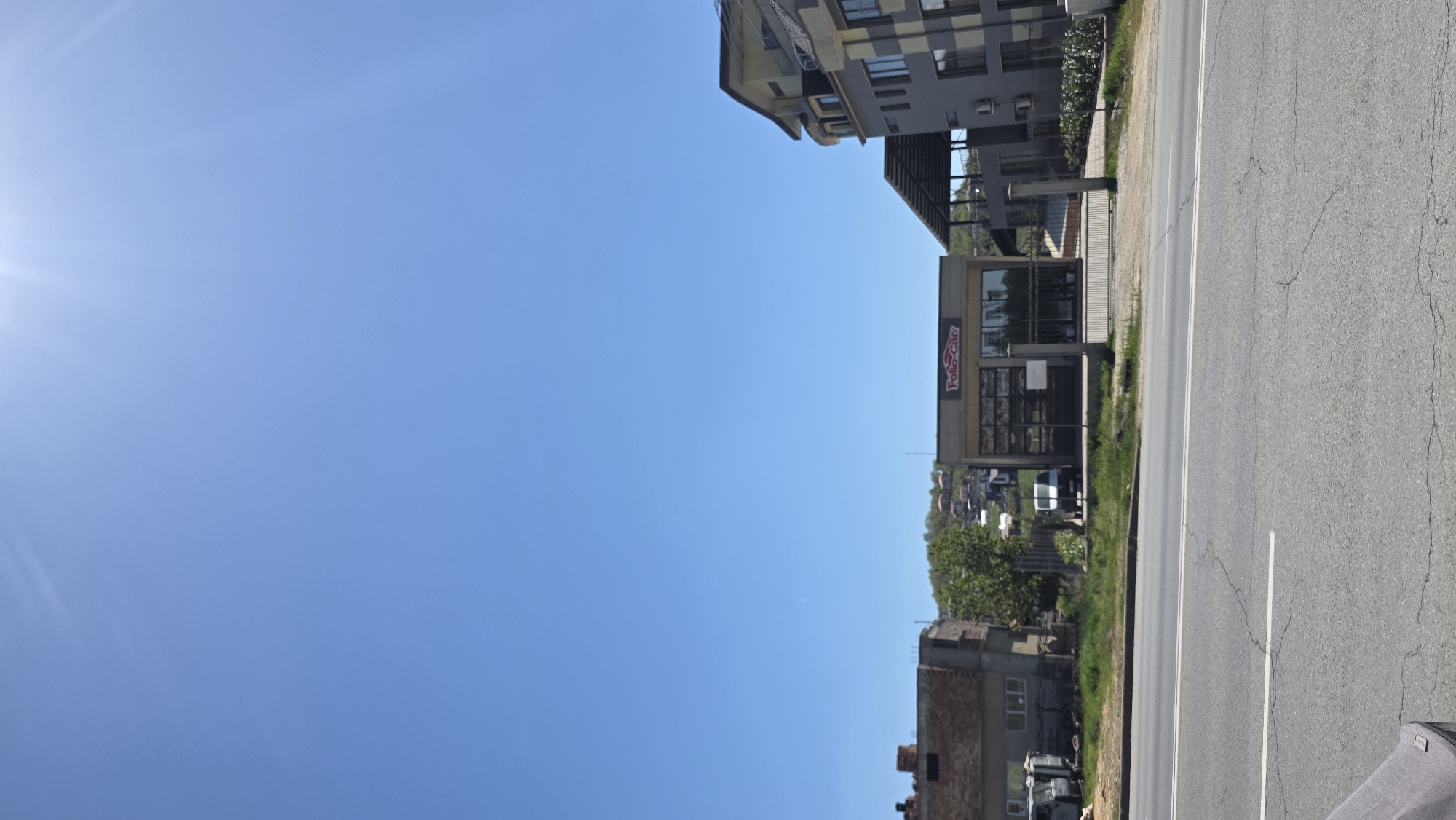 The image shows a street scene in Blagoevgrad, Bulgaria. The sky is clear and blue. A building is located to the right side of the image, including a business named "Folk Car". A road and the side of a building are also visible on the right side of the image.