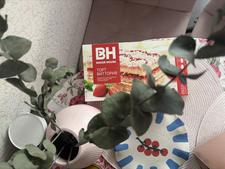 The image depicts an indoor scene in Novosaratovka, Russia, viewed from an overhead perspective. In the foreground, a leafy green branch, consistent with eucalyptus, extends from a light pink, spirally ribbed ceramic vase. A white Nutella brand lid is partially visible behind and to the left of the vase. The central item is a rectangular box for "ТОРТ ВИТТОРИЯ" (Vittoria Cake) by "BAKER HOUSE," predominantly red and white, displaying an image of a layered strawberry cake. To the lower left of the cake box, an irregularly shaped white ceramic plate or coaster is decorated with red berries on a green stem and several blue linear segments along its edge. These objects rest on a light-colored, possibly woven, placemat. Beneath and surrounding these items, a light-colored fabric with a red and pink floral pattern is visible. To the right of the cake box, a smooth, light pink, upholstered surface is present. The background shows a light-colored wall with a subtle textured pattern. No individuals are present, and no actions are depicted.