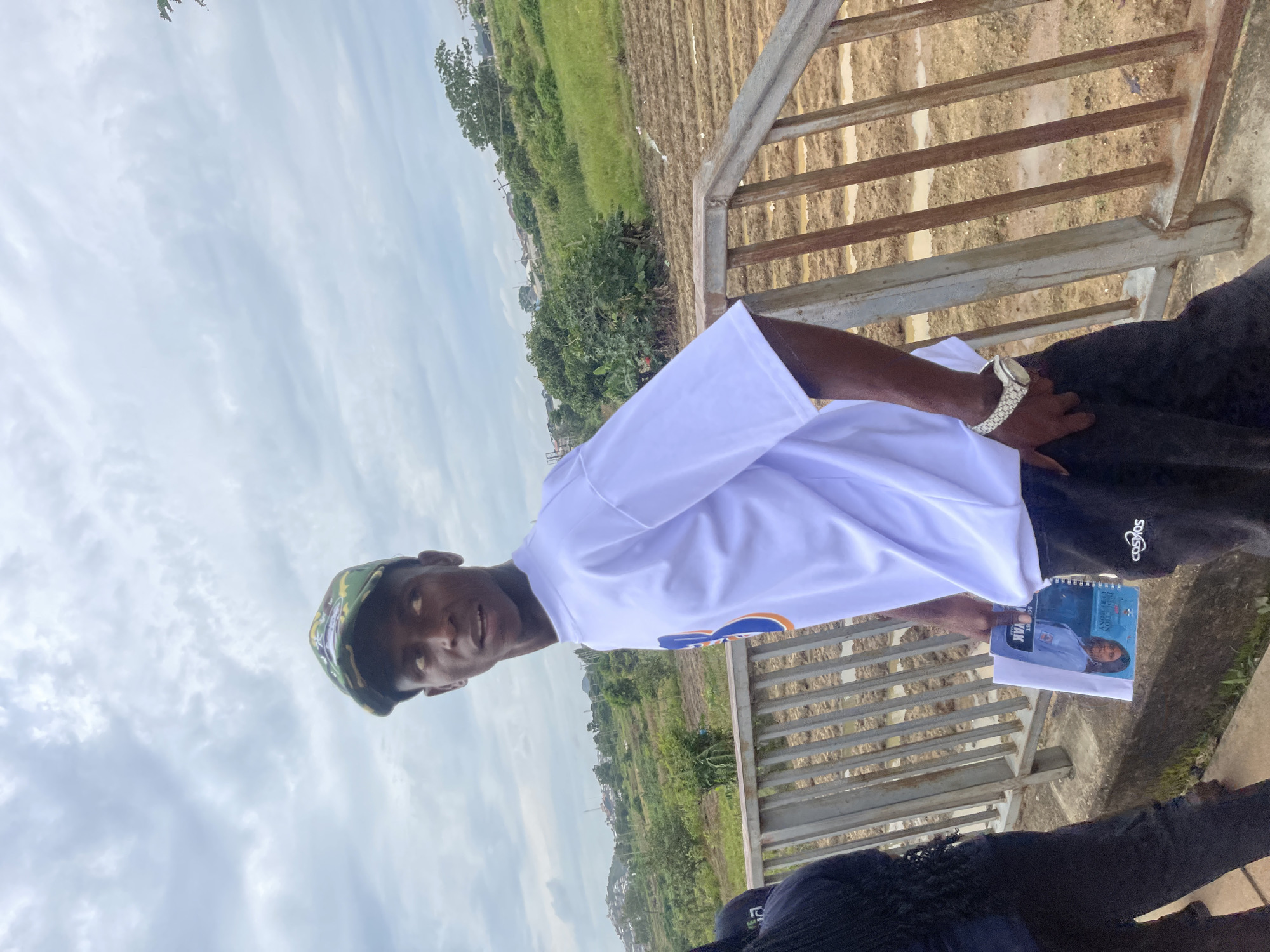 The image depicts a young man standing on a pedestrian bridge, looking away from the camera. He is wearing a camouflage-patterned baseball cap, a white t-shirt with an orange and blue graphic on the sleeve, and black sweatpants with a white "Cosmos" logo. He is holding a notebook or binder in his left hand. To his left, another person wearing black clothing is partially visible. The bridge has metal railings. Beyond the bridge, there are fields with rows of what appear to be crops or tilled soil, some of which are wet, suggesting recent rain. In the background, there are trees and buildings, indicating a semi-urban or rural setting. The sky is overcast with gray clouds. The location is identified as Rafin Par, Nigeria.