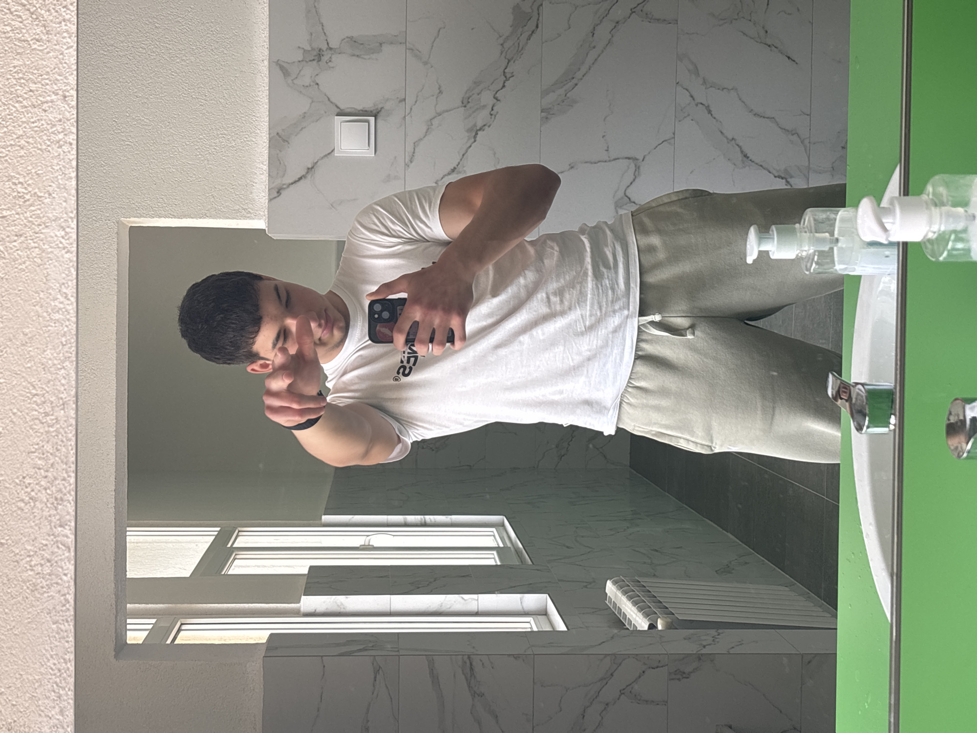 A young man is taking a selfie in a bathroom, likely in Zlatograd, Bulgaria. He is wearing a white t-shirt and grey sweatpants, holding a black smartphone with a case. He is pointing with his left hand, as if mimicking a gun. The bathroom features white marble-patterned walls, a sink with soap dispensers, a green-framed mirror reflecting the scene, and a window with light entering. A white radiator is visible on the floor.