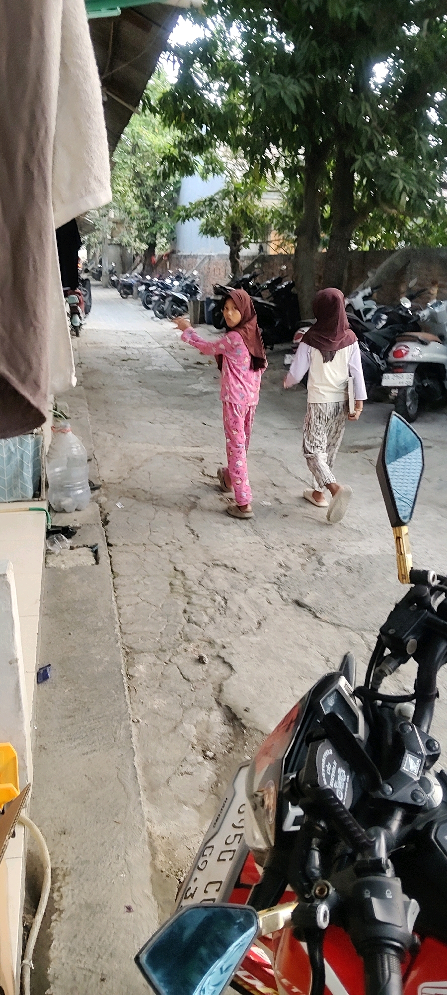 Two young girls, wearing hijabs and modest clothing, are walking down a paved alley in Mekarmukti, Indonesia. The girl in the foreground on the left is wearing pink pajamas with a floral pattern and a brown hijab. She is pointing her left arm towards the distance. The girl on the right is wearing a white top, patterned pants, and a brown hijab, and she is holding a book or notepad in her right hand. In the background, several motorcycles are parked on both sides of the alley. Trees with green foliage are visible overhead, and a portion of a building with a blue wall is present. In the foreground, a motorcycle is partially visible, with its handlebars, mirrors, and license plate in view. The license plate appears to be white with black text. The ground is cracked and uneven. There is a large plastic water bottle on the left side of the alley, near a low concrete wall.