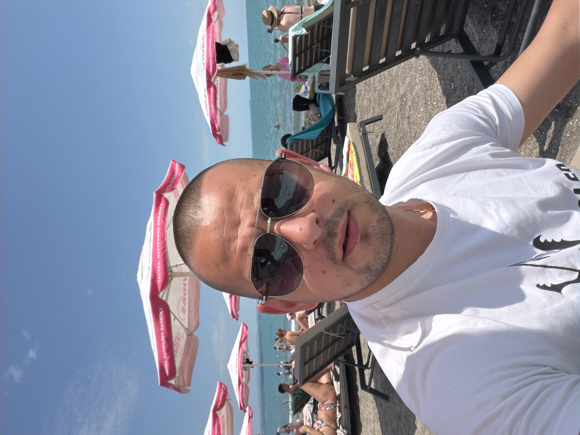 A close-up, eye-level outdoor shot depicts a man at a bustling beach in Burgas, Bulgaria, under a clear blue sky. The foreground is dominated by the man, who has a buzzed haircut, light stubble, and wears dark aviator-style sunglasses reflecting the scene. He is wearing a white crew-neck t-shirt with a dark graphic partially visible on the lower left and a gold chain around his neck; his expression is neutral with a slightly parted mouth.
The midground and background reveal a sun-drenched beach with dark sand or fine gravel. Multiple beachgoers are visible, including adults and children, some reclining on dark-framed lounge chairs with colorful towels (blue, yellow, green), others walking, or swimming in the calm, blue-green sea. Several large white beach umbrellas with prominent pink/red trim are scattered across the beach, bearing Cyrillic text such as "ЧЕСТНА, НАТУРАЛНА НАПИТКА" (Honest, Natural Drink) and "VIDAS" (reflected on some). The horizon line where the sea meets the sky is clearly visible.