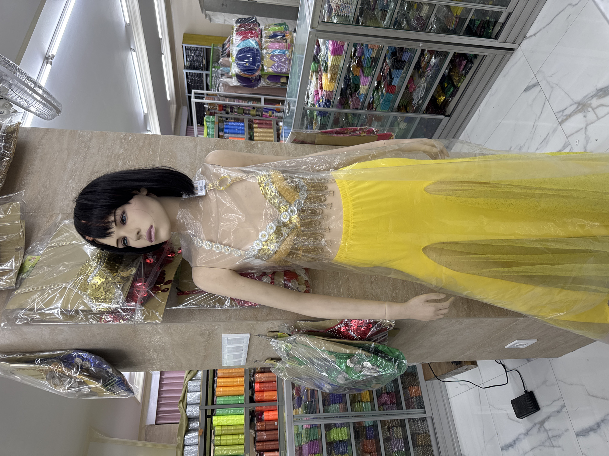 A mannequin dressed in a yellow belly-dancing outfit is laying on a shelf in a store. The mannequin has a black bob wig and is covered in plastic wrap. The top of the outfit is white with gold sequins and tassels. The bottom is a long, flowing yellow skirt. The shelf is made of beige stone. In the background, there are shelves stocked with various colorful textiles. The store is brightly lit with fluorescent lights. The location is likely in Kuta, Indonesia.