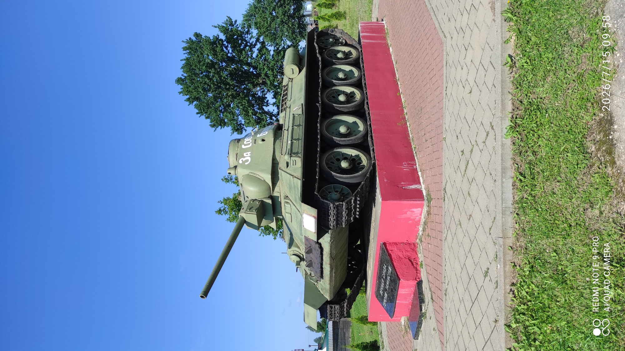 A historical Soviet T-34 tank, painted olive green, is displayed on a red concrete pedestal. The tank is positioned on a paved area surrounded by brickwork and grass. Behind the tank, there are trees, including a large, mature deciduous tree. The sky is clear blue. A black plaque is visible on the front of the pedestal, bearing an inscription. The scene is captured on a bright, sunny day.