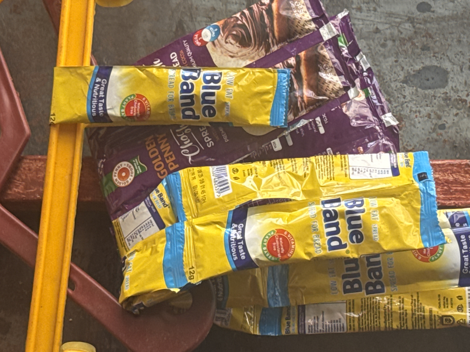 This media file displays several packages of Blue Band spread and Golden Penny spread arranged on a surface, possibly a table or shelf. The products are primarily yellow and purple, with clear branding and nutritional information visible on the packaging. The yellow packages feature the "Blue Band" logo prominently, along with text such as "Great Taste & Nutritious" and "Low Fat Spread." Some packages also highlight "6 VITAMINS" and "PLANT BASED." The purple packages, identified as "GOLDEN PENNY SPREAD," show images of chocolate swirls and text indicating "CHOCOLATE" and "PREMIUM QUALITY."
The overall scene appears to be a product display or storage area, with a blurred background that includes elements of a textured surface and some reddish-brown and yellow objects, possibly part of furniture or shelving. The lighting suggests an indoor setting during daylight hours. There are no people visible in the image. The arrangement of the products suggests they are either for sale or have been recently acquired. The location context of Abuja, Nigeria, provides a geographical setting for these commonly available food products in the region. The focus is entirely on the product packaging, emphasizing their visual design and branding.