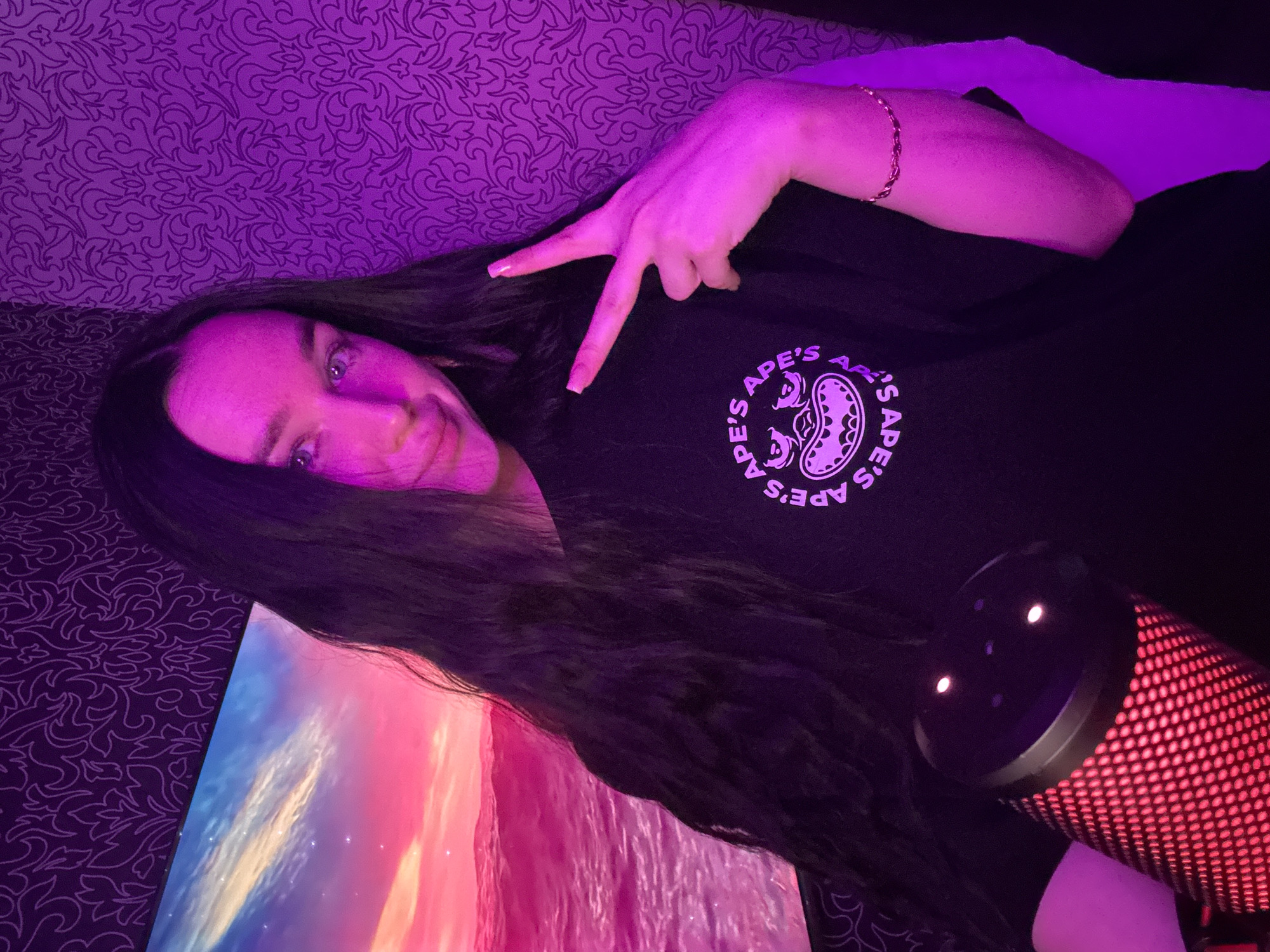 A young woman with long dark hair is pictured from the chest up, indoors. She is wearing a black t-shirt with a circular logo featuring a cartoonish face. The woman is making a peace sign with her right hand. To her left, a portion of a computer monitor displaying a sunset over water is visible. In the foreground on the left, a red microphone with a mesh cover is present. The background consists of purple patterned wallpaper. The scene is lit with purple and pink lights. The image does not contain enough information to determine the specific location in Novosibirsk, Russia.