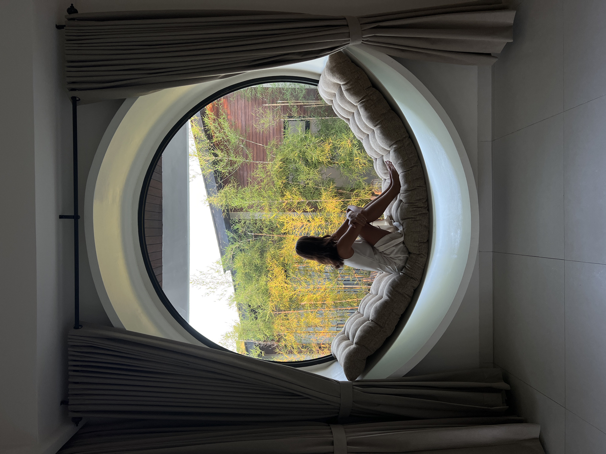 A woman sits on a cushioned window seat, looking out at a bamboo garden through a large, circular window. The window is framed by two beige curtains. The interior is sparsely furnished with tiled flooring. The bamboo garden appears well-maintained, with a building visible behind it. The scene suggests a peaceful and contemplative atmosphere. The image is likely taken in a residential or hospitality setting in Ubud District, Indonesia, given the architectural style and tropical vegetation.