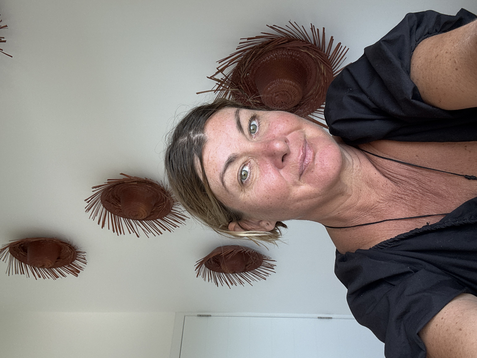 A medium-shot portrait captures a woman indoors, her head tilted to the right, looking directly at the camera with a faint, friendly smile. She has light brown hair, green eyes, and visible freckles on her face. A thin black cord necklace is visible on her neck, which also shows some fine wrinkles. She is wearing a black top or dress with gathered fabric on the shoulder.
The background consists of a plain white wall or ceiling, adorned with several decorative, woven straw objects. These items resemble small, flat straw hats or stylized sunbursts with radiating fringes, colored in shades of brown. One large straw decoration is positioned above and behind her head, while three smaller ones are arranged on the wall to the left. A glimpse of a white door or doorframe is visible in the lower right portion of the image.
The setting appears to be a well-lit indoor space with a simple, decorative aesthetic. The style of the straw decorations could suggest a tropical or rustic influence, which aligns with the provided location context of Baan Chaweng Noi, Thailand. The woman's expression is calm and serene. There is no visible text in the image.