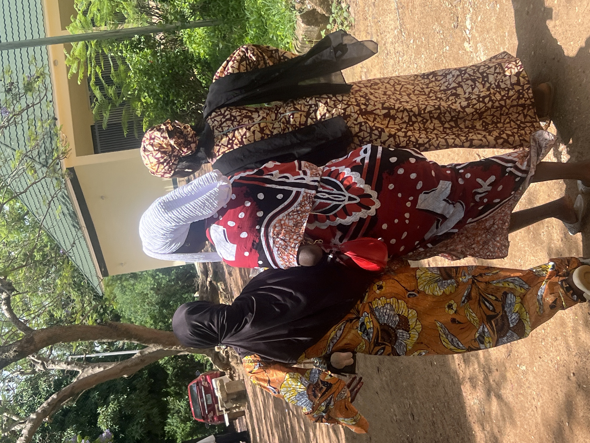 Three women dressed in patterned fabrics and head coverings are walking on a sunlit, concrete surface. The woman in the foreground wears a black head covering and a patterned orange and brown dress. The second woman wears a white head covering and a patterned red and white dress. The third woman wears a brown patterned head covering with a black scarf and a patterned brown dress. Trees and vegetation are visible in the background, along with a building. A red vehicle is visible behind the women. The location is Jos, Nigeria.