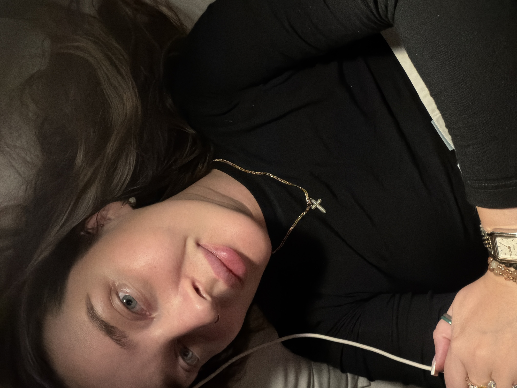 The image captures a person, likely a woman, lying down indoors, looking directly at the camera. The perspective is rotated, with her head in the lower-left portion of the frame and her body extending towards the upper-right. She has long, dark brown hair fanned out around her and light blue eyes.
She is dressed in a black long-sleeved top. A gold chain necklace with a small, crystal-embellished cross pendant hangs around her neck. Her right wrist is adorned with a rectangular-faced wristwatch and several beaded bracelets. Multiple rings are visible on her fingers, including one with a green stone and another with several lighter-colored stones. A small stud or hoop nose piercing is present on her left nostril, and a subtle earring is visible in her left ear.
The background suggests an indoor setting, possibly a bed or couch, with soft, light-colored fabric. The dim lighting implies an evening or nighttime scene. A white cable, resembling a headphone or charger cord, stretches across the lower part of the image, near her right hand. Her facial expression is neutral and calm. No visible text is present in the image. The location context provided is Moscow, Russia.