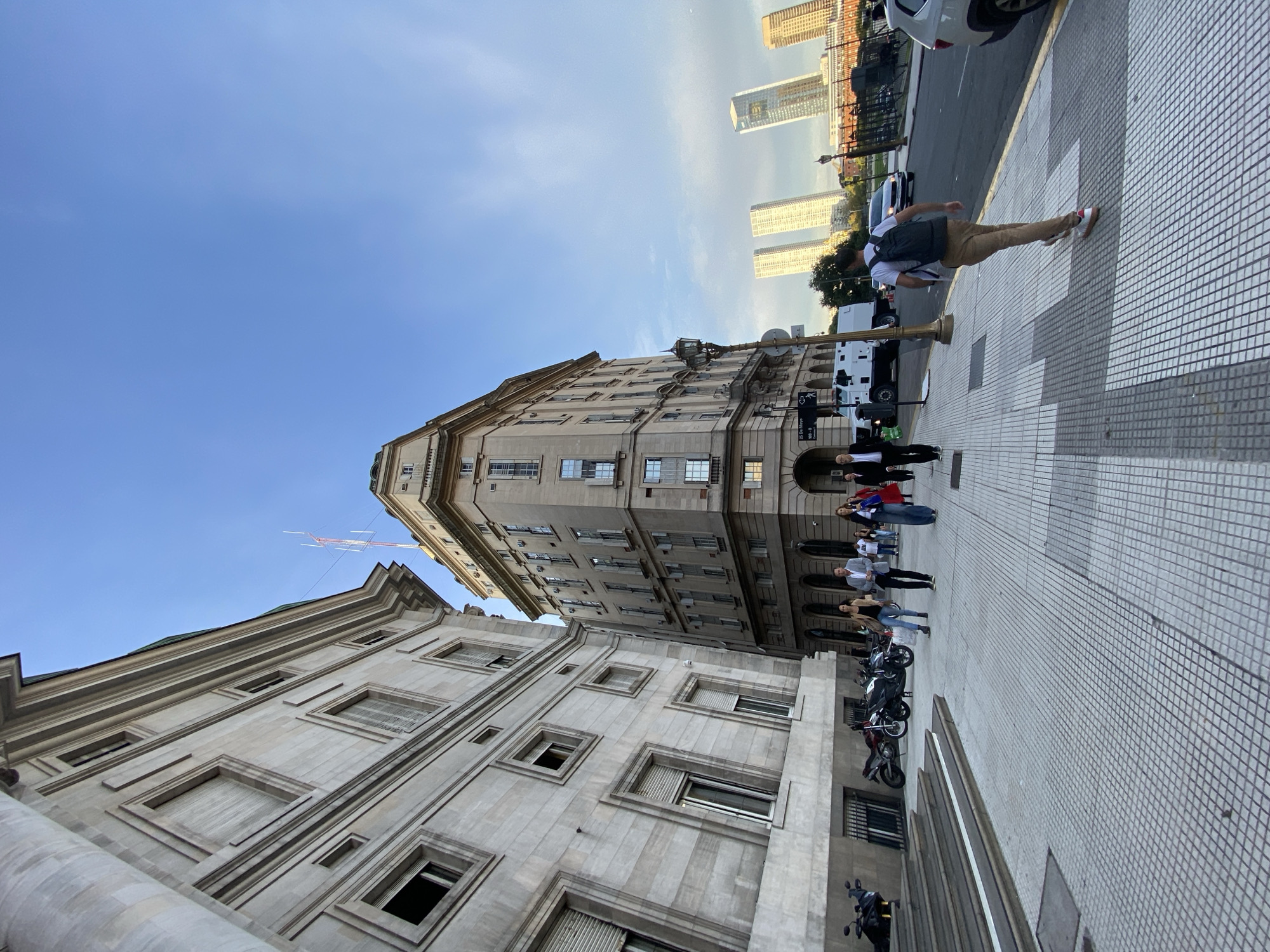 The image shows a street scene in Buenos Aires, Argentina. The camera angle is tilted. Tall, classical-style buildings flank the street. A group of people stands on the sidewalk. There are also parked motorcycles. In the distance, modern skyscrapers are visible. A pedestrian is crossing the street. A truck is parked near the sidewalk. The sky is blue.