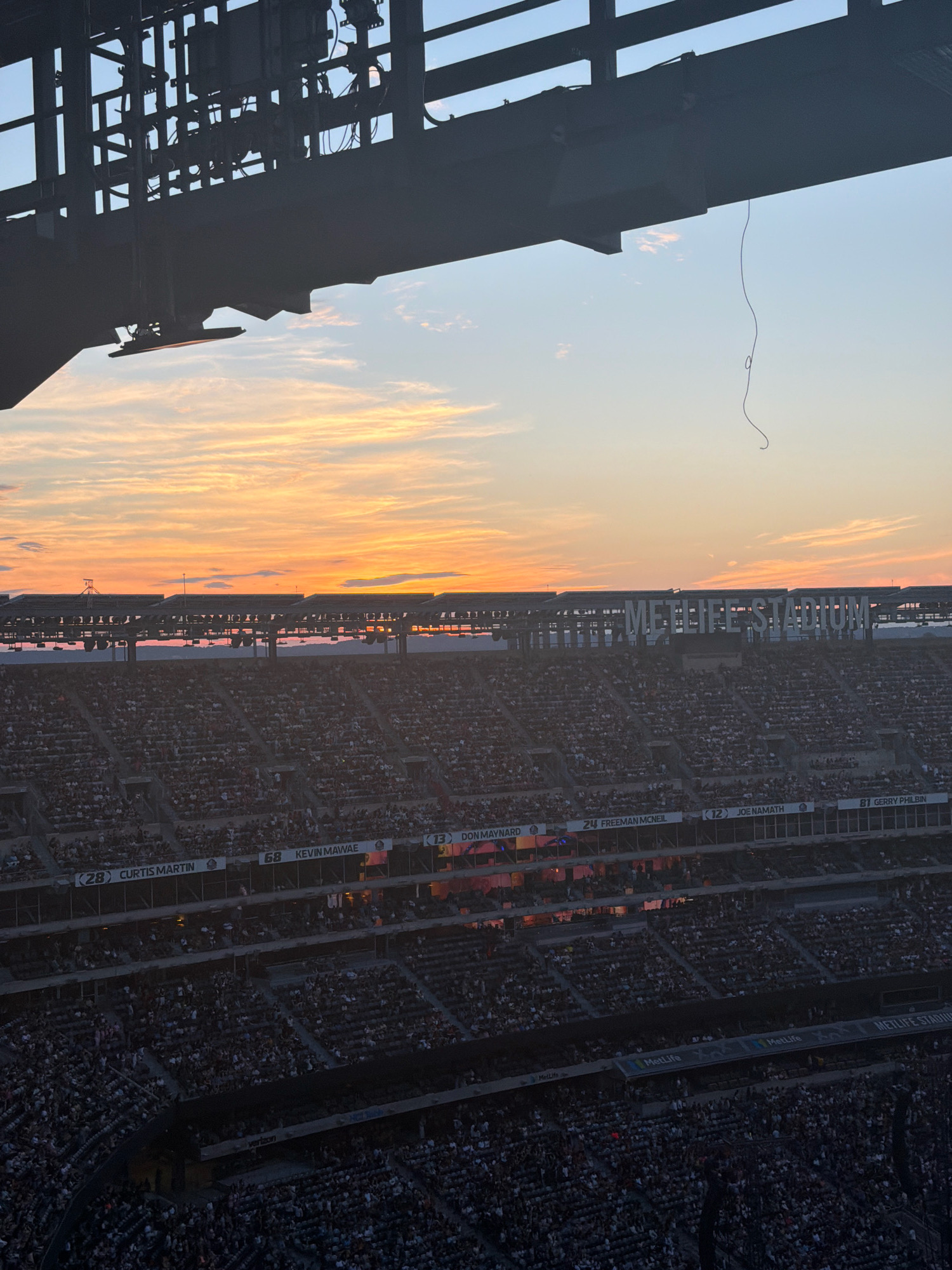 This image depicts an exterior view of MetLife Stadium in East Rutherford, New Jersey, United States of America, during sunset. The sky is filled with warm hues of orange and pink from the setting sun, with scattered clouds. A large, dark metal structure, likely part of the stadium's infrastructure or upper seating, dominates the foreground and top portion of the image. A thin, dark wire hangs from this structure.
Below this, rows upon rows of stadium seating are visible, populated by a large crowd of spectators. The lower sections of the stadium are darker, suggesting the sun is behind the stadium from this perspective. The name "METLIFE STADIUM" is prominently displayed in large white lettering on the stadium's upper deck. Various banners with numbers and names of athletes, such as "CURTIS MARTIN," "KEVIN MAWAE," "DON MAYNARD," "FREEMAN MCNEIL," "JOE NAMATH," and "GERRY PHILBIN," are visible on different levels of the stadium. The overall scene captures a bustling atmosphere at a sports venue during an event, set against a picturesque sunset.
