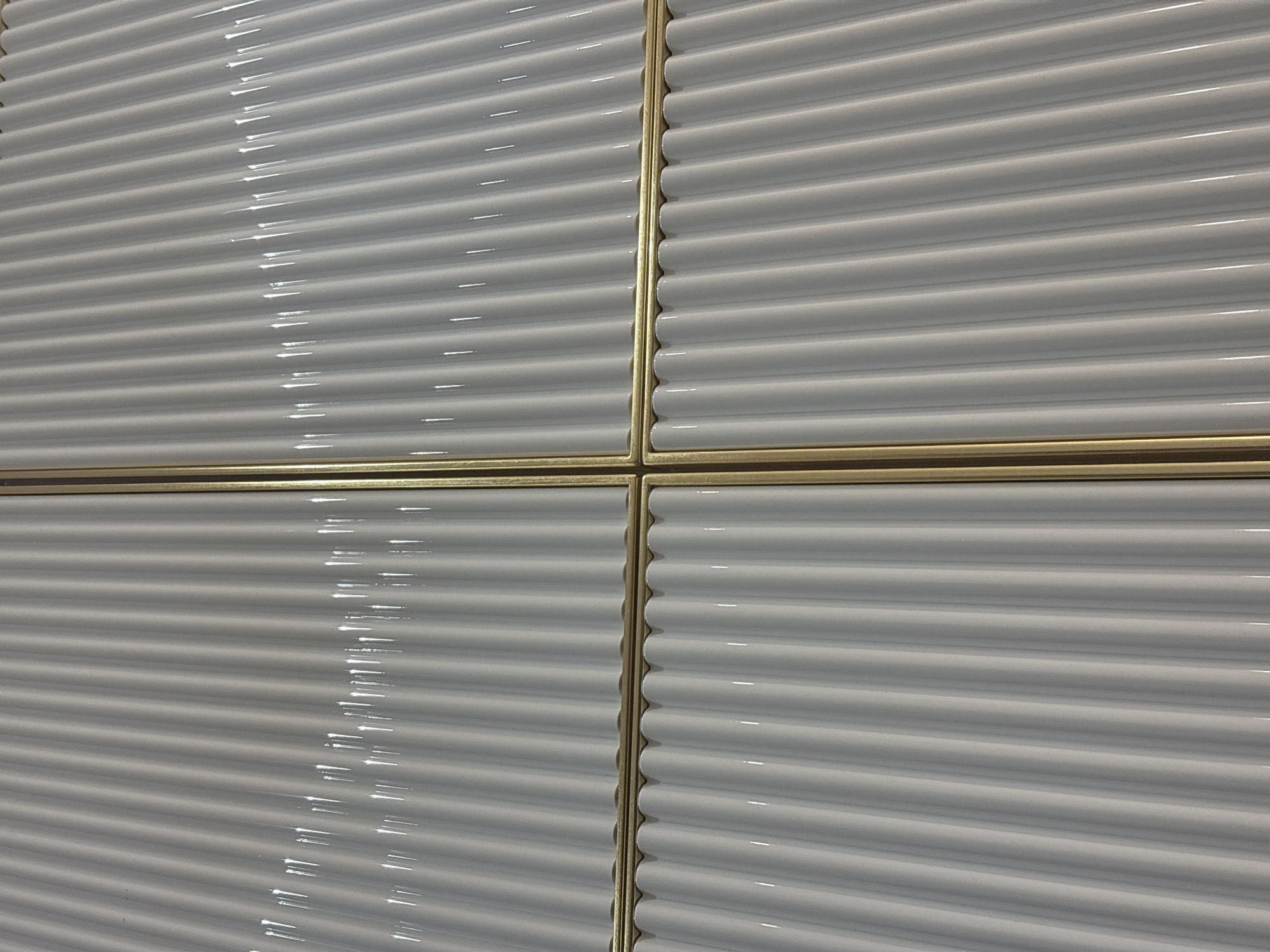 The image shows a close-up of a tiled wall. The tiles are white with a corrugated, rounded texture, arranged horizontally. Thin, gold-colored metal trim separates the tiles. The lighting is bright, creating highlights on the glossy tile surfaces. The scene depicts a modern interior design element. There are no people present.