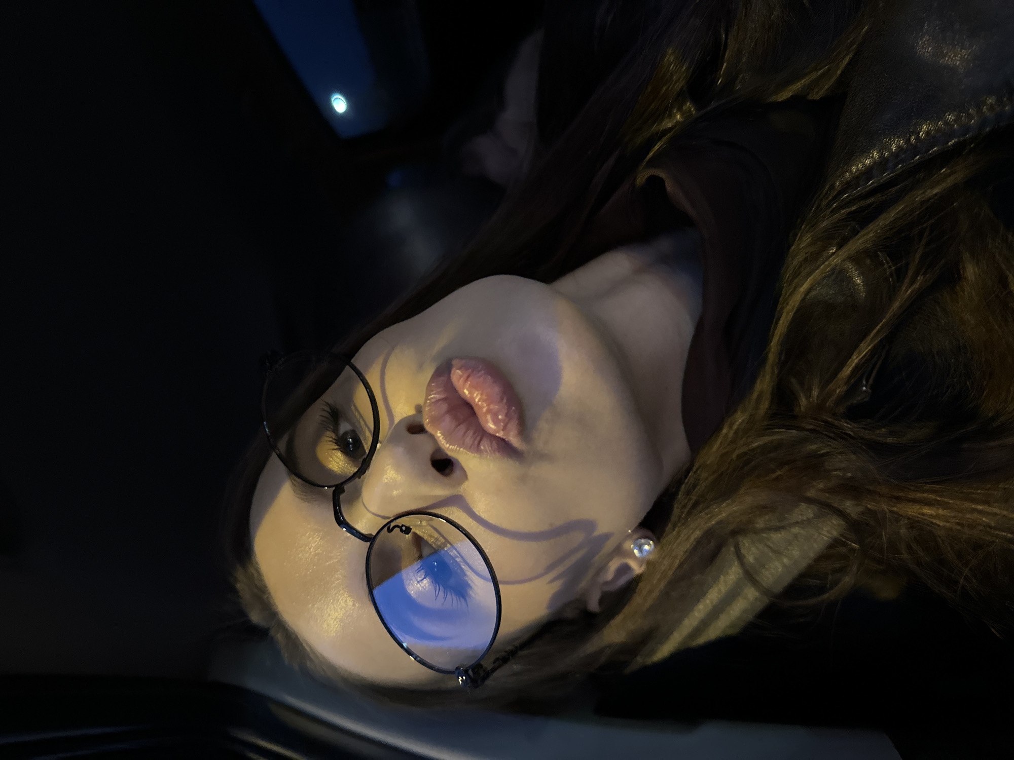 A portrait-style photograph captures a woman indoors, presumably in a car at night. The photo is oriented such that the woman's face is angled, with her head tilted to the left. She wears round glasses reflecting an external blue light source. The woman has full lips and an earring in her ear. Her dark hair is partially visible, alongside a leather jacket. The environment is dark, with a hint of an interior car surface. The city is not discernible.