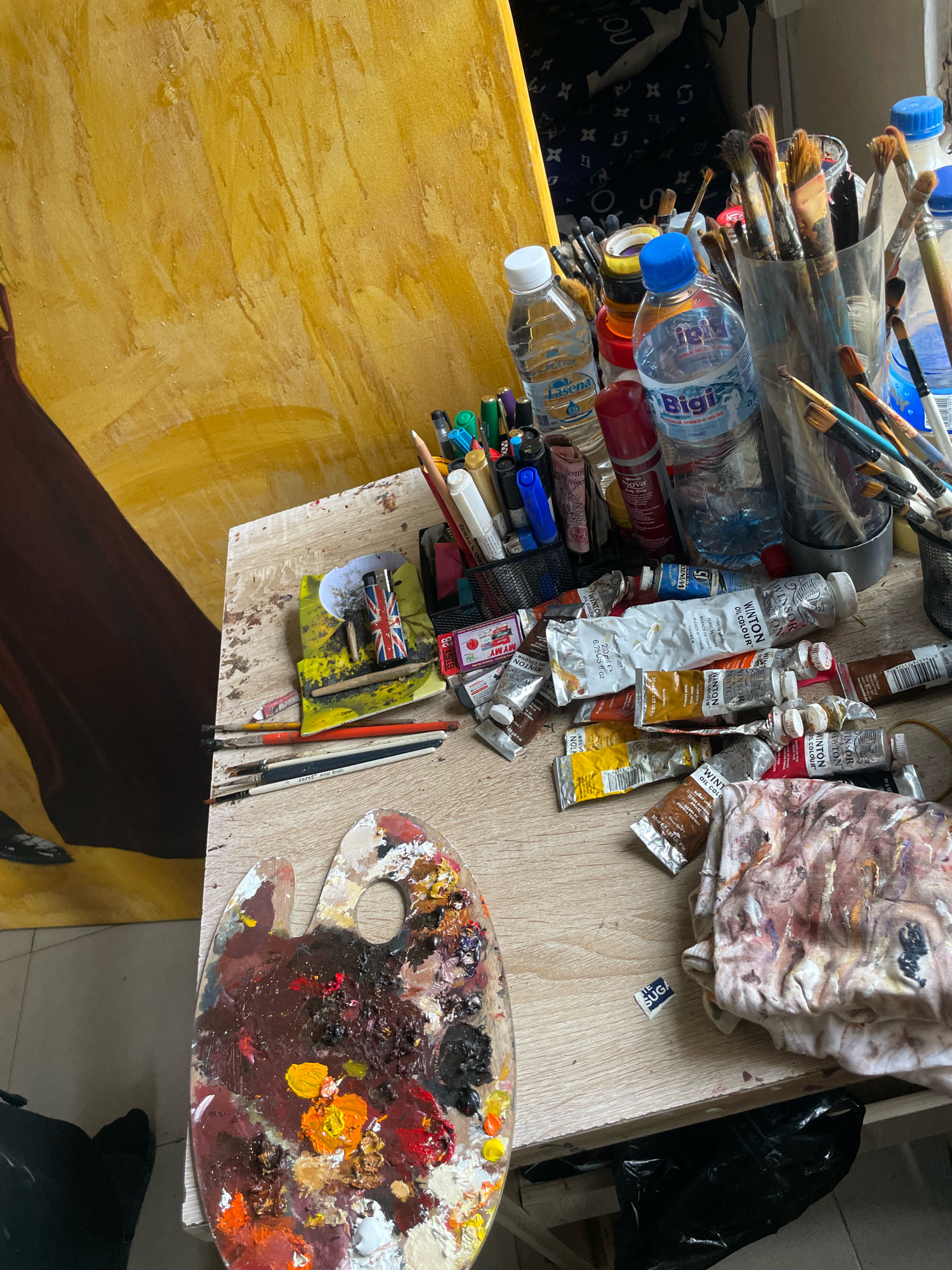 An artist's cluttered wooden table in Ado, Nigeria, is the setting for this still life. A large, unfinished yellow canvas dominates the background, with a figure in a dark red garment partially visible on its left side. The table is covered with an assortment of art supplies. In the foreground, a wooden palette is laden with a vibrant array of oil paints in shades of brown, red, yellow, and white. Scattered across the table are numerous tubes of oil paint, primarily from the "Winton Oil Colour" brand. Several plastic water bottles, two with blue caps and one with a white cap, are interspersed with the paints and other items. Among the art supplies, a metal cup holds a collection of paintbrushes of various sizes. A small, folded cloth, smudged with paint, lies near the bottom right of the frame. Other objects on the table include a lighter adorned with the Union Jack flag, a small packet labeled "SUG," and several pens and pencils. The overall impression is of a working studio, filled with the tools and materials of an artist's craft.