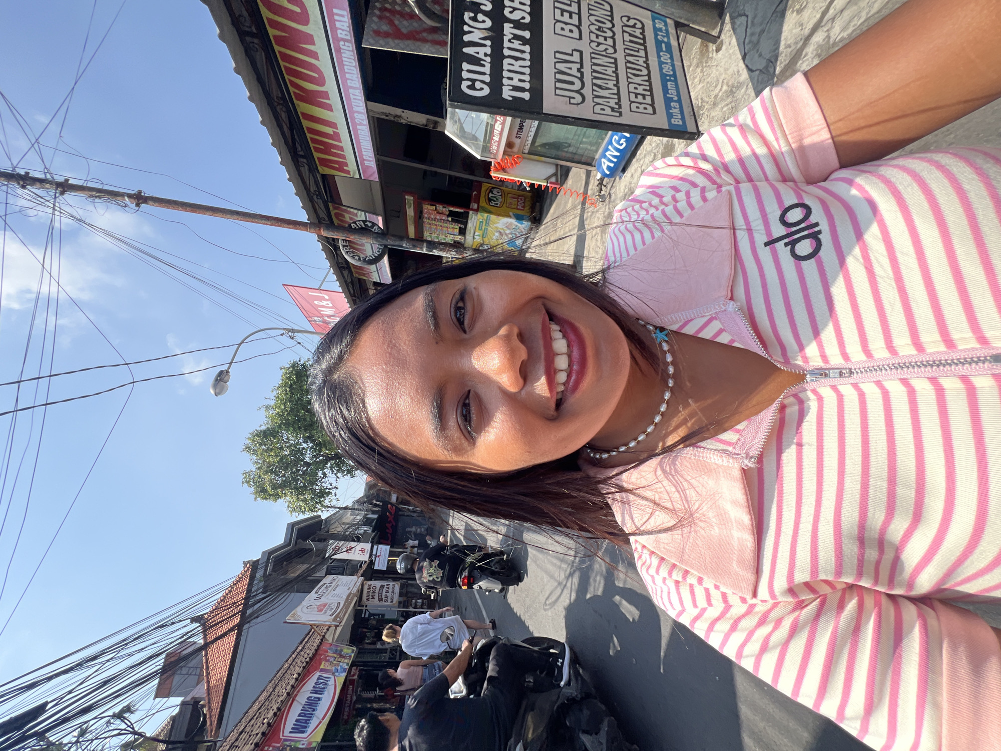 A woman stands in the middle of a bustling street in Kuta, Indonesia, taking a selfie. She has dark hair, brown eyes, and is smiling broadly, revealing her teeth. She is wearing a pink and white striped collared zip-up shirt with the logo "alo" on the left side of her chest. A pearl necklace with a blue star pendant is around her neck.
The street is lined with shops, signs, and an abundance of overhead electrical wires. To the left, a red and white sign reads "WARUNG HESTI". Further down, another sign displays "WARUNG MIKO". A woman in a white shirt and a man on a motorcycle are visible in the background. To the right, a dark sign with white text reads "GILANG JAYAKARTA THRIFT SHOP" with additional Indonesian text below that, indicating business hours. The road is paved and reflects the sunlight. The sky above is clear and blue. The overall atmosphere suggests a warm, sunny day in a commercial area of Kuta.