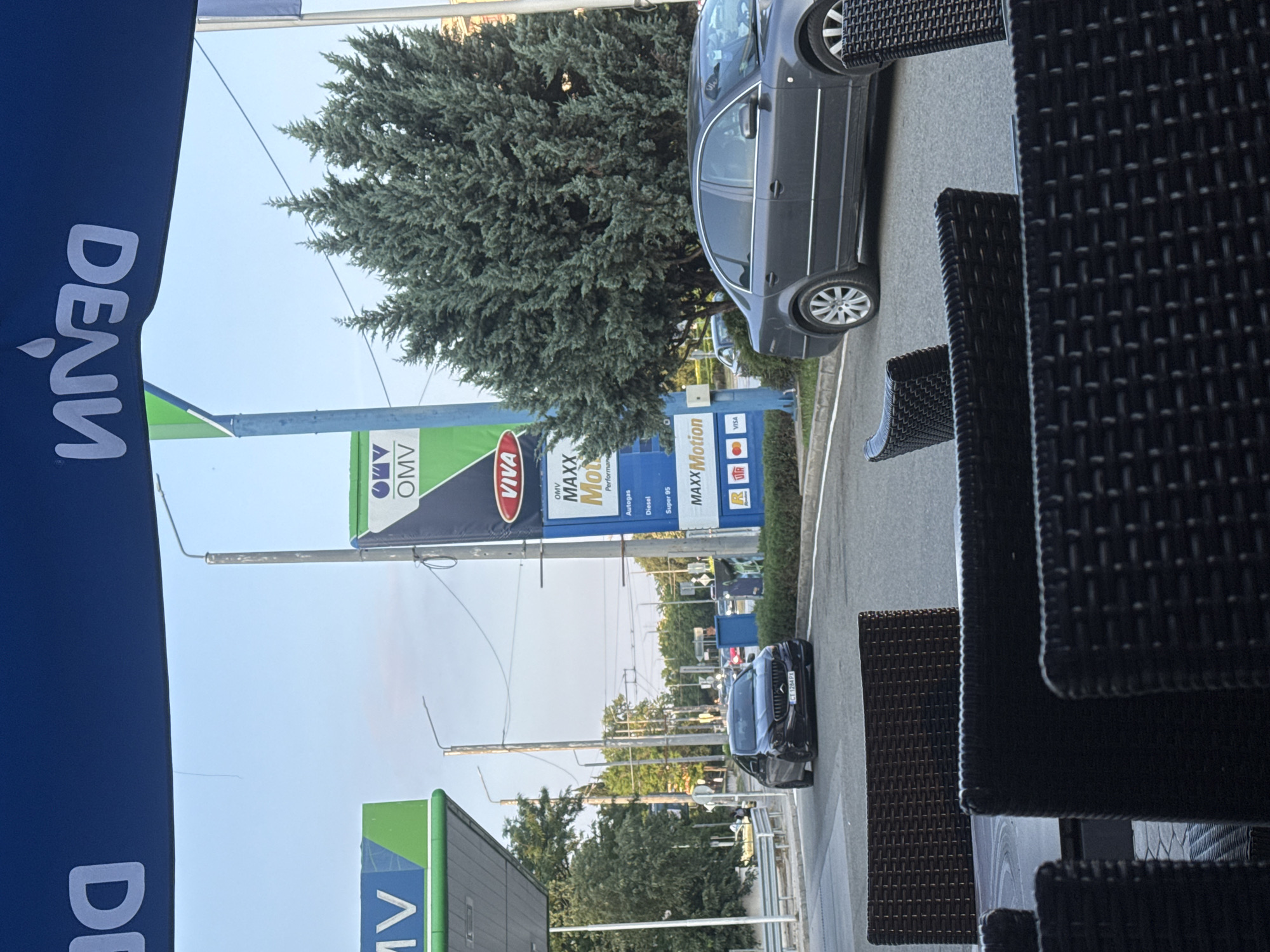 The image depicts an outdoor scene in Stara Zagora, Bulgaria, specifically at an OMV gas station and an adjacent street. The vantage point is from beneath a blue awning or banner, which displays the reversed white text "DENIM" across the top.
In the immediate foreground, dark brown, woven outdoor seating furniture (chairs and table sections) is partially visible, indicating the photograph was taken from a cafe or restaurant patio.
The midground features an asphalt road with traffic. On the left, a black sedan, identified by its Bulgarian license plate "CT 1284 PX" (Stara Zagora region), is visible turning or positioned on the road. On the right, a dark grey sedan is parked or stationary. A prominent, large evergreen tree with dense foliage stands between the road and the gas station's pricing totem.
Central to the midground is an OMV gas station. A tall, blue and green OMV totem displays fuel information, including "MAXXMotion," "Autogas," "Diesel," and "Super 95," along with payment logos such as "Mastercard" and "VISA." Another OMV sign is partially visible on the far left, under a gas station canopy structure. Overhead streetlights/utility poles are also visible along the roadside.
The background shows additional trees and faint outlines of distant buildings under a clear, light blue sky. No individuals are clearly discernible in the image.