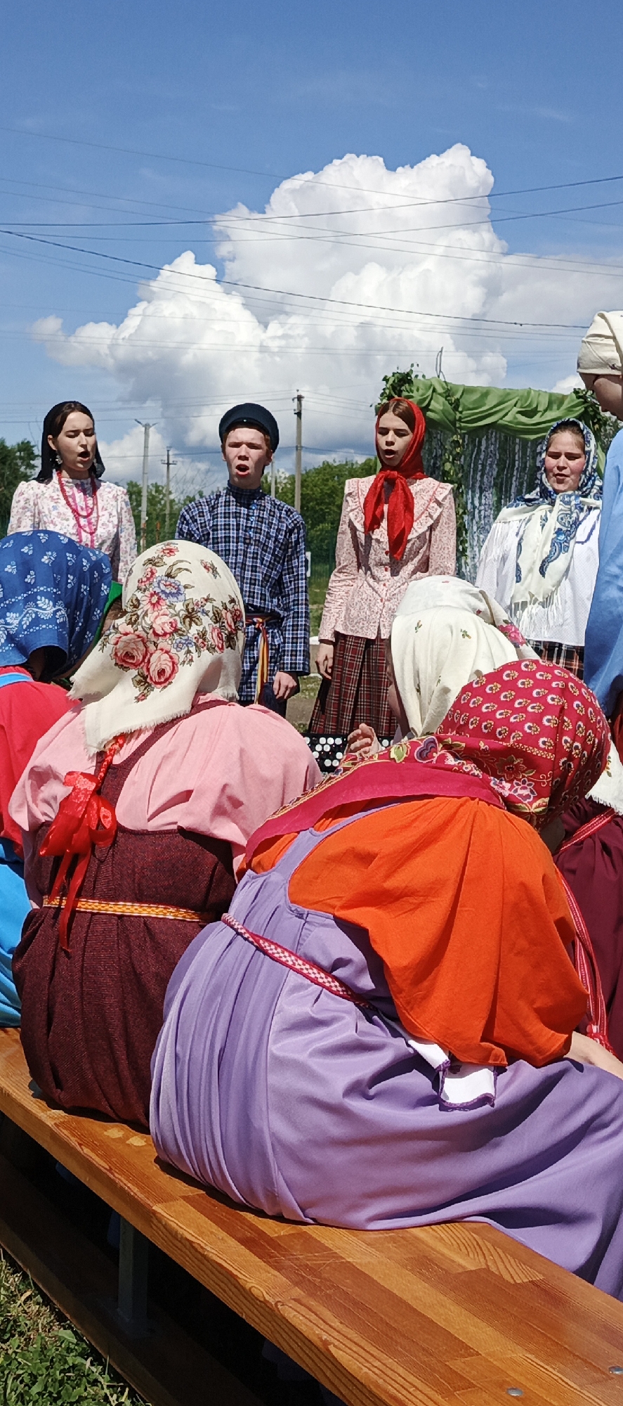 The photograph depicts a group of people gathered in what appears to be a performance setting in the city of Кемуль, Russia. In the foreground, several people are seated on a wooden bench, their backs to the camera. They wear traditional Russian clothing, including brightly colored dresses and headscarves with floral patterns.
Behind the seated individuals, a small group of people stands and faces the camera. They also wear traditional Russian clothing. The backdrop includes trees, power lines, and a large white cloud in a blue sky. The overall setting is outdoors, and the photograph appears to have been taken during a sunny day.