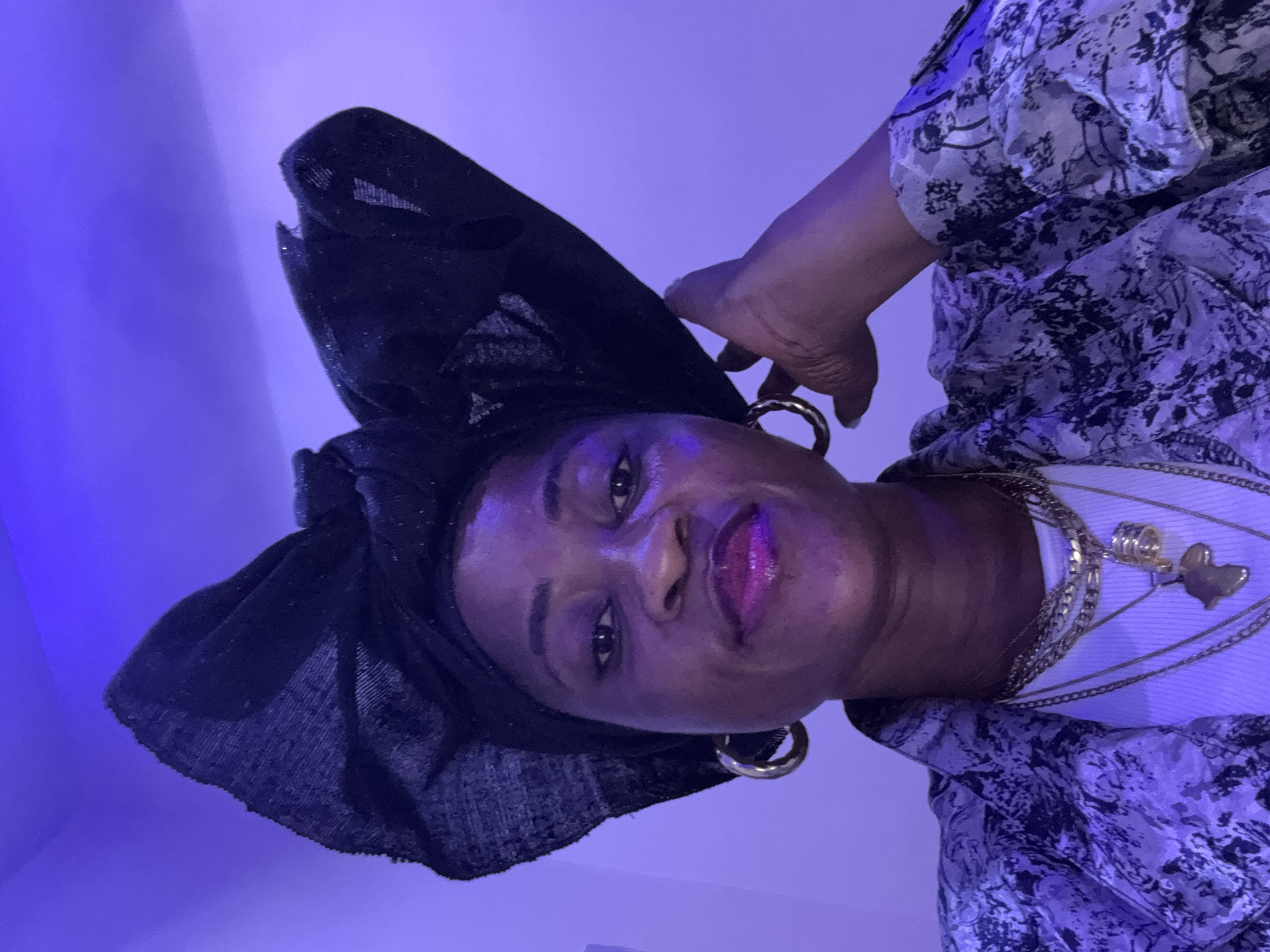 A self-portrait of a woman, adorned in a black headwrap with a large bow, gold hoop earrings, and a patterned jacket over a white top, is captured in a dimly lit room with purple lighting. Her hand is resting on her headwrap. The background is a plain, light-colored wall. The image does not provide any specific details about the city of Ajara, Nigeria, nor does it offer any context for the woman's location within it.