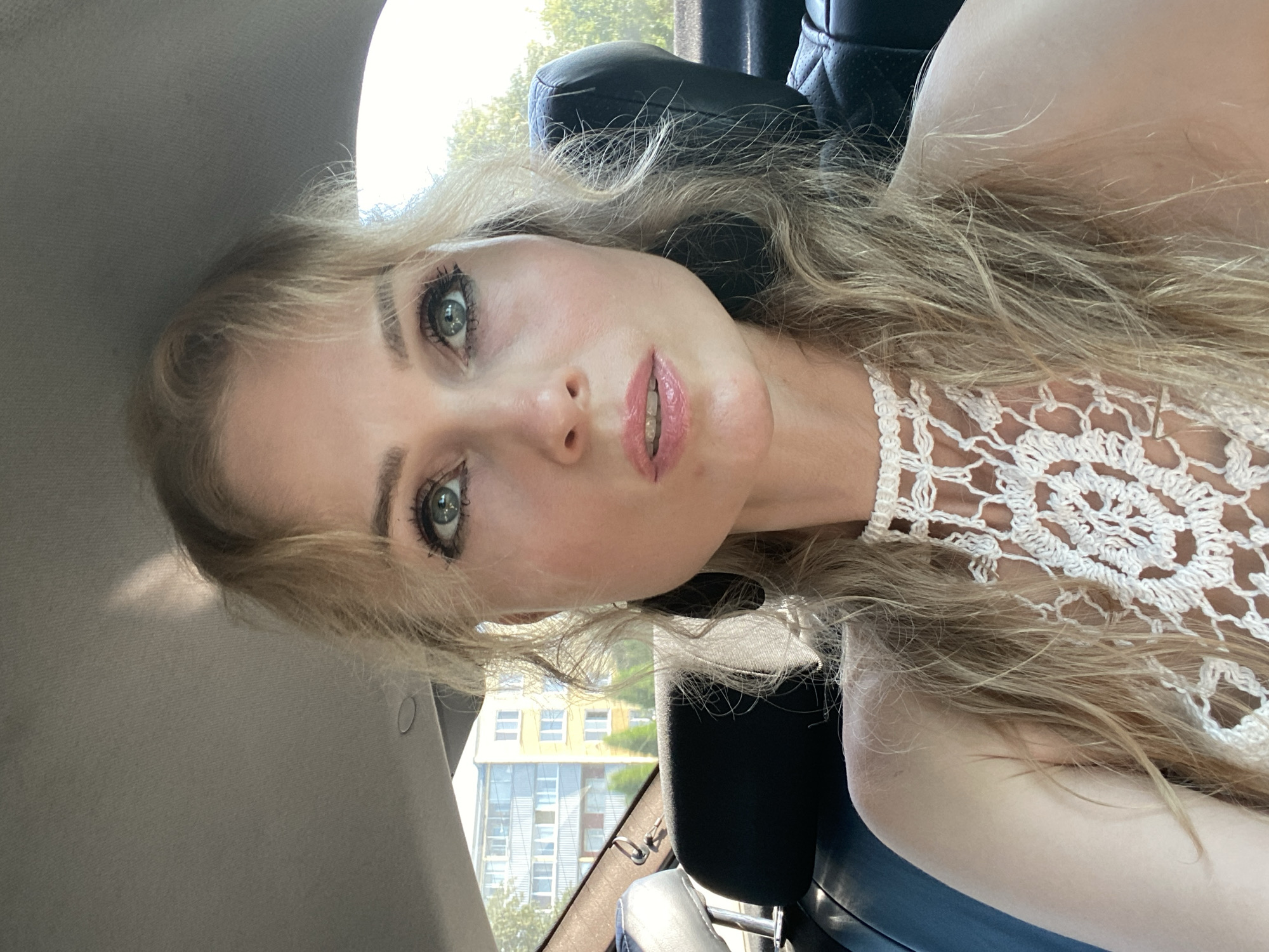 A close-up selfie shows a blonde woman with blue eyes and light makeup sitting in the back seat of a car. She is wearing a white crocheted top. The background shows buildings and trees, suggesting an urban or suburban setting. The car interior includes black leather seats and the ceiling of the car.