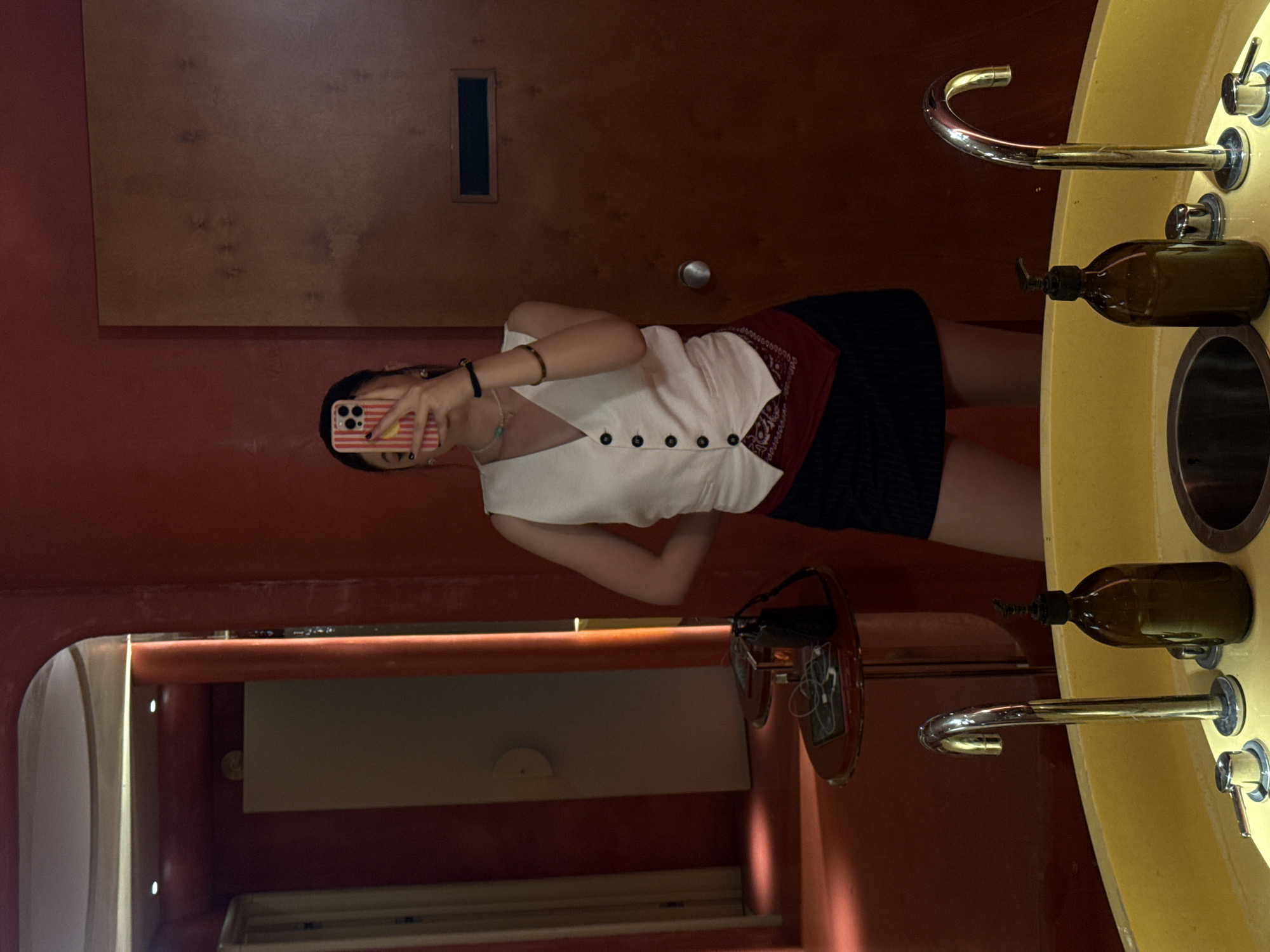 The image displays an indoor mirror selfie, likely taken in a restroom or a changing area. The location, identified as Yerevan, Armenia, is not explicitly visible within the photograph itself.
A single individual, a female, is centered in the frame, holding a mobile phone to obscure her face while capturing her reflection. She wears a cream-colored, sleeveless, V-neck vest with six black buttons down the front, a dark-colored mini-skirt, and a red bandana or fabric piece tucked into her waistband on her left side. Her visible accessories include a multi-strand beaded necklace in various colors (white, turquoise), a black elastic bracelet on her left wrist, and a brown bangle on her right arm. Her phone features a distinctive red and white striped case with a small yellow sticker.
In the immediate foreground, a bright yellow, circular or oval vanity unit with a built-in dark drain is visible. Two polished metal faucets are installed on the yellow surface; one is a tall, arched gooseneck style on the right, and the other is a shorter, lever-operated faucet on the left. Two amber-colored glass pump bottles, likely containing soap or lotion, are positioned on the vanity.
To the left of the individual, a small, round table with a dark red, reflective surface stands on a metallic pedestal base. On this table, a black power bank, white wired earbuds, and a small electronic device (possibly a tablet or phone) are present.
The room's walls are a uniform, textured reddish-orange. To the left, an arched entryway reveals a lighter-colored door (possibly off-white or light gray) that is partially ajar, with recessed ceiling lights visible beyond the arch. To the right of the individual, a plain, dark brown wooden door with a round silver doorknob is visible. This door also features a rectangular black vent or recess near its top. The lighting in the room is warm and dim, highlighting the red and yellow color scheme, with a prominent vertical light strip illuminating the inner side of the arched passage on the left. The floor appears to be consistent with the reddish-orange wall color.
