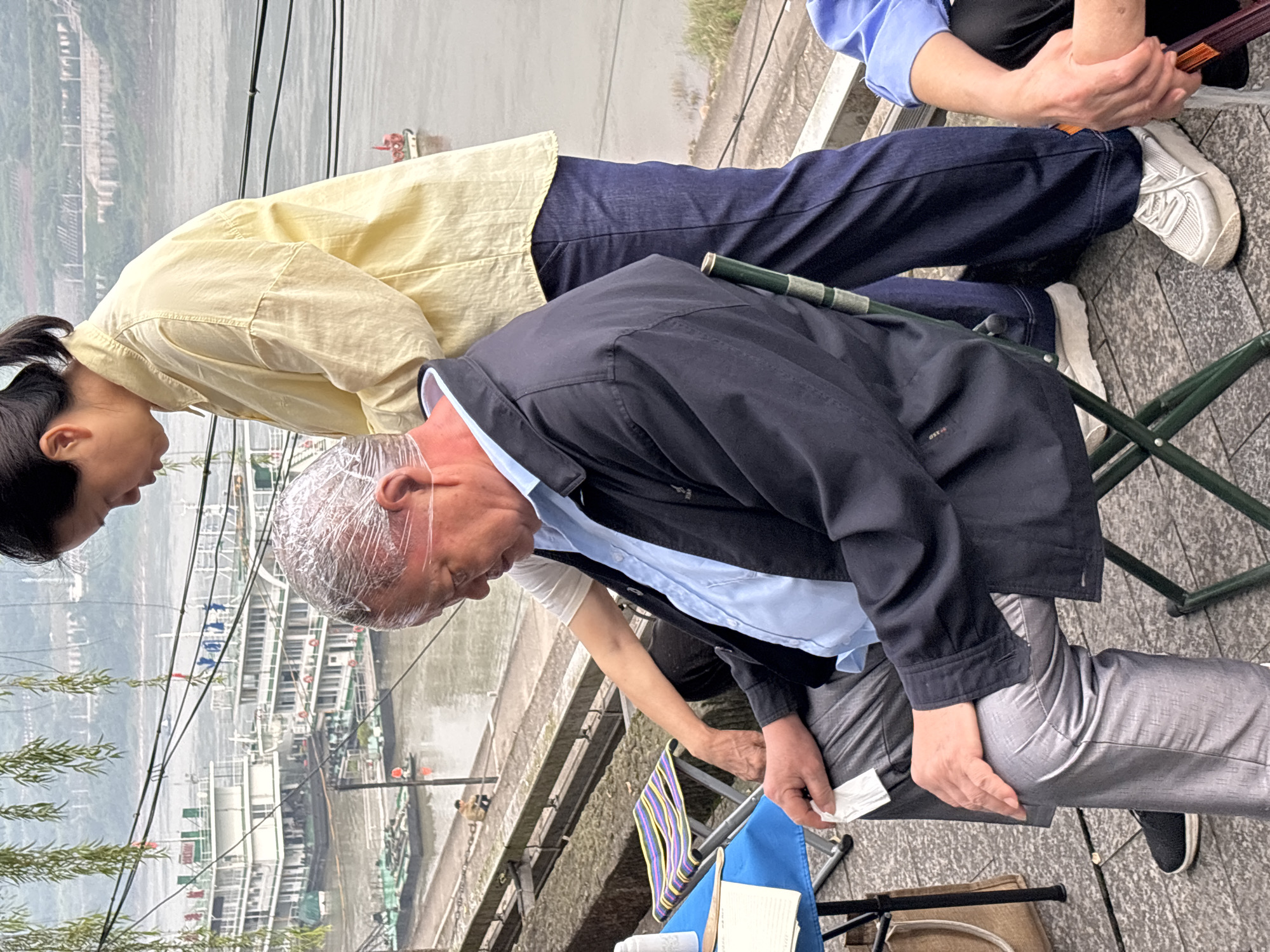 In Jiangjin, China, an elderly man with his head wrapped in plastic film sits on a folding chair by a river. A woman in a yellow shirt stands behind him, and another woman's arms are visible in the foreground. A table with a blue cloth and a blue chair are also present. In the background, several boats are docked on the river, and hills can be seen on the far bank. The scene appears to be outdoors, possibly in a park or along a promenade, with a paved walkway leading to the river.