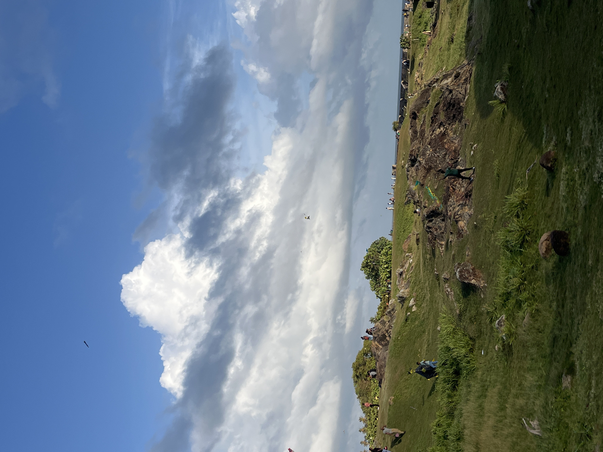 The image depicts a wide, grassy hillside dotted with rocks and a few trees, overlooking a body of water, likely the ocean. The sky is a mix of blue with dramatic white and grey clouds, suggesting it might be late afternoon or early evening. Several people are scattered across the hillside, some walking, others sitting or standing. A notable activity is kite flying, with multiple kites visible in the sky and some individuals actively engaged in flying them. The landscape is typical of coastal areas, with rolling green terrain and rocky outcrops. In the distance, the outline of a structure, possibly an old fort, can be seen along the coastline. The overall impression is one of a public space, likely a park or a recreational area, where people are enjoying an outdoor activity with a scenic view. Given the mention of Galle, Sri Lanka, this scene could be within the vicinity of Galle Fort, a historic architectural heritage site known for its ramparts and scenic coastal views.
