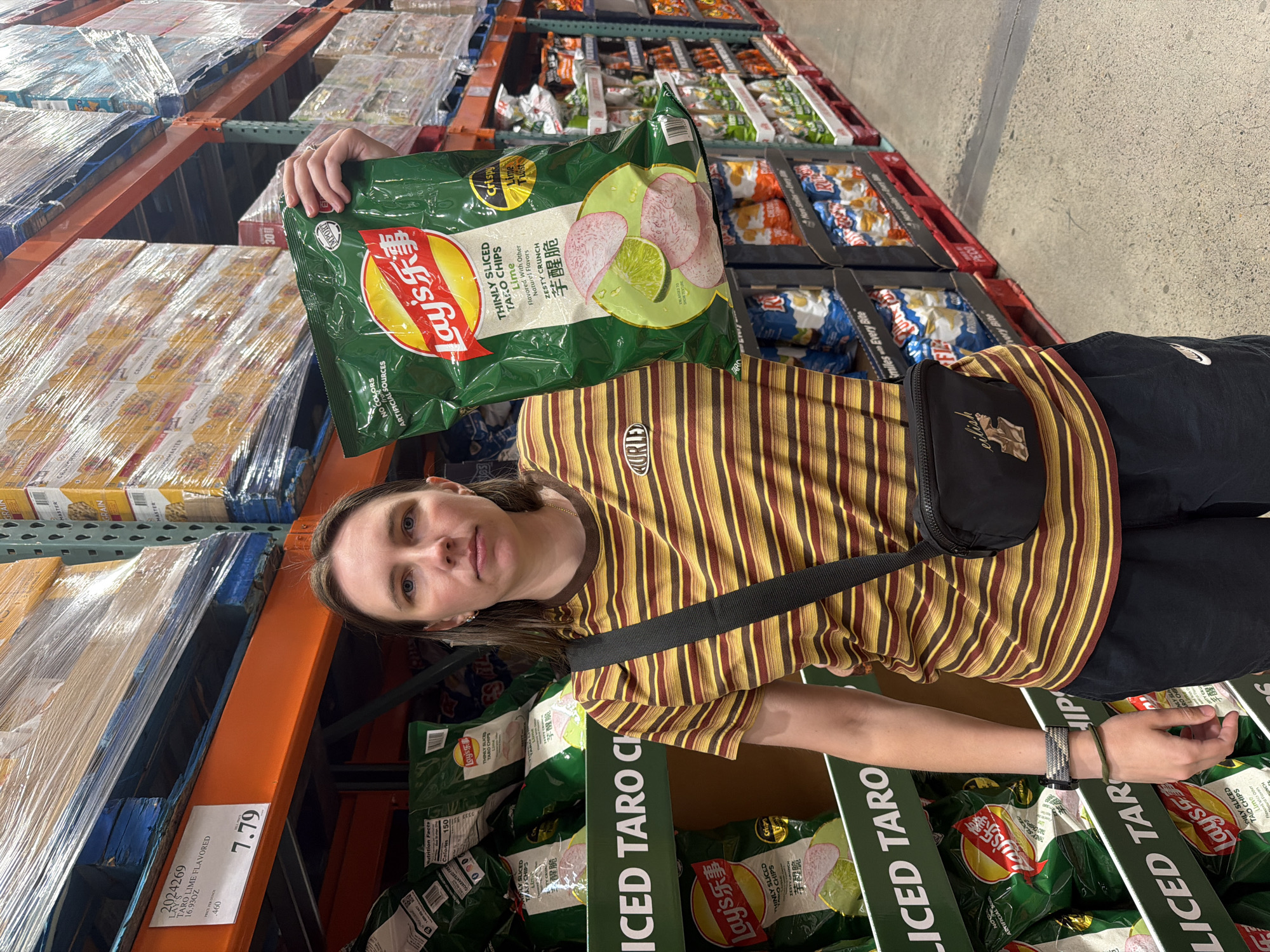 In Beaverton, Oregon, a woman stands in a Costco store, holding a large bag of Lay's thinly sliced taro chips with lime flavor. She is wearing a horizontally striped t-shirt in shades of brown and yellow, black shorts, and a black fanny pack with a graphic detail. She has on a watch and a bracelet on her left wrist. In the background, shelves are stocked with various chips and food products. The store environment appears to be a typical warehouse-style retail setting.
