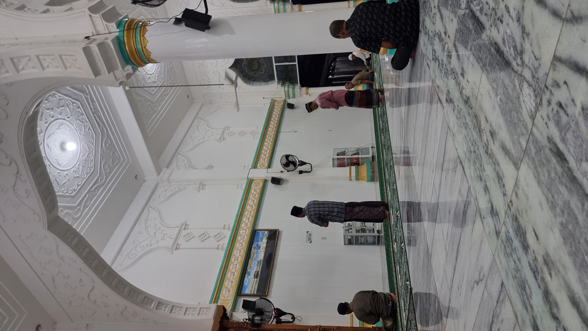 The image depicts the interior of a mosque in Lapang Timu, Indonesia, characterized by its clean, white walls adorned with intricate moldings and a domed ceiling with a light fixture. The floor is made of polished marble with a reflective surface, and green prayer rugs are laid out. Several men are present, engaged in prayer or observing. One man in the foreground is sitting on the floor facing away from the camera, wearing a black shirt with a repeating pattern and black pants. Another man stands in the middle ground, wearing a plaid shirt and a sarong, facing a framed picture on the wall. To the right, two men are seen in prayer, one wearing a pinkish tunic and sarong, and another in a light brown shirt and sarong. In the background, more men are visible, some kneeling on prayer mats. The mosque's architecture includes arched doorways, white columns with gold and turquoise accents, and ceiling fans. Wall-mounted fans and a picture of the Kaaba are also visible. The overall atmosphere suggests a moment of quiet devotion within the mosque.