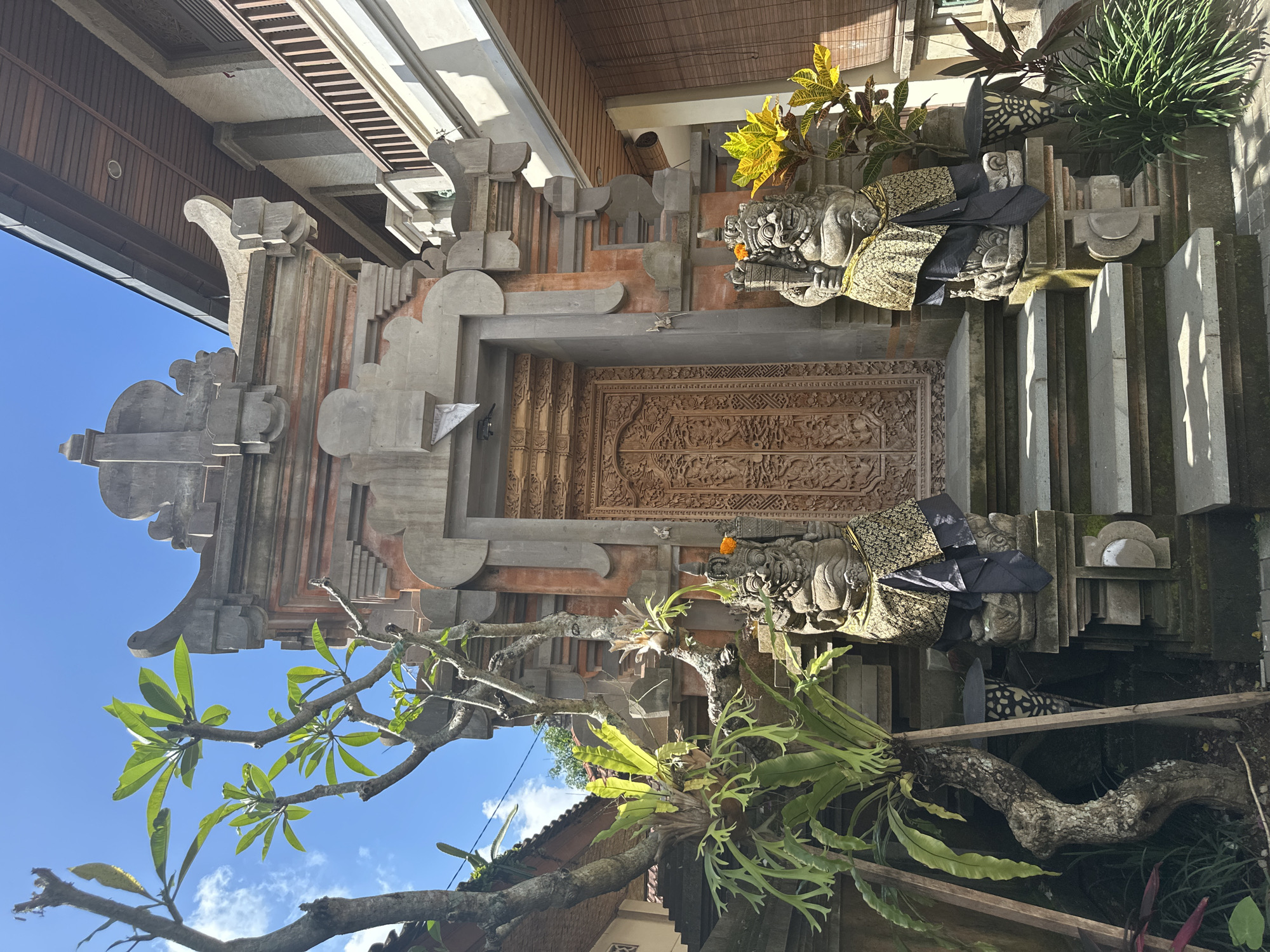 The image depicts the entrance to a traditional Balinese building, likely a temple or a house, located in the Ubud District of Indonesia. The architecture features intricate stone carvings and ornate wooden doors. Flanking the entrance are two guardian statues, adorned with traditional Balinese patterned cloths. Lush green foliage, including a tree with broad leaves and hanging ferns, surrounds the entrance, adding to the tropical ambiance. The sky above is clear blue with scattered white clouds. The scene is brightly lit by natural sunlight, casting shadows on the steps leading up to the doorway. The overall impression is one of cultural richness and natural beauty.
