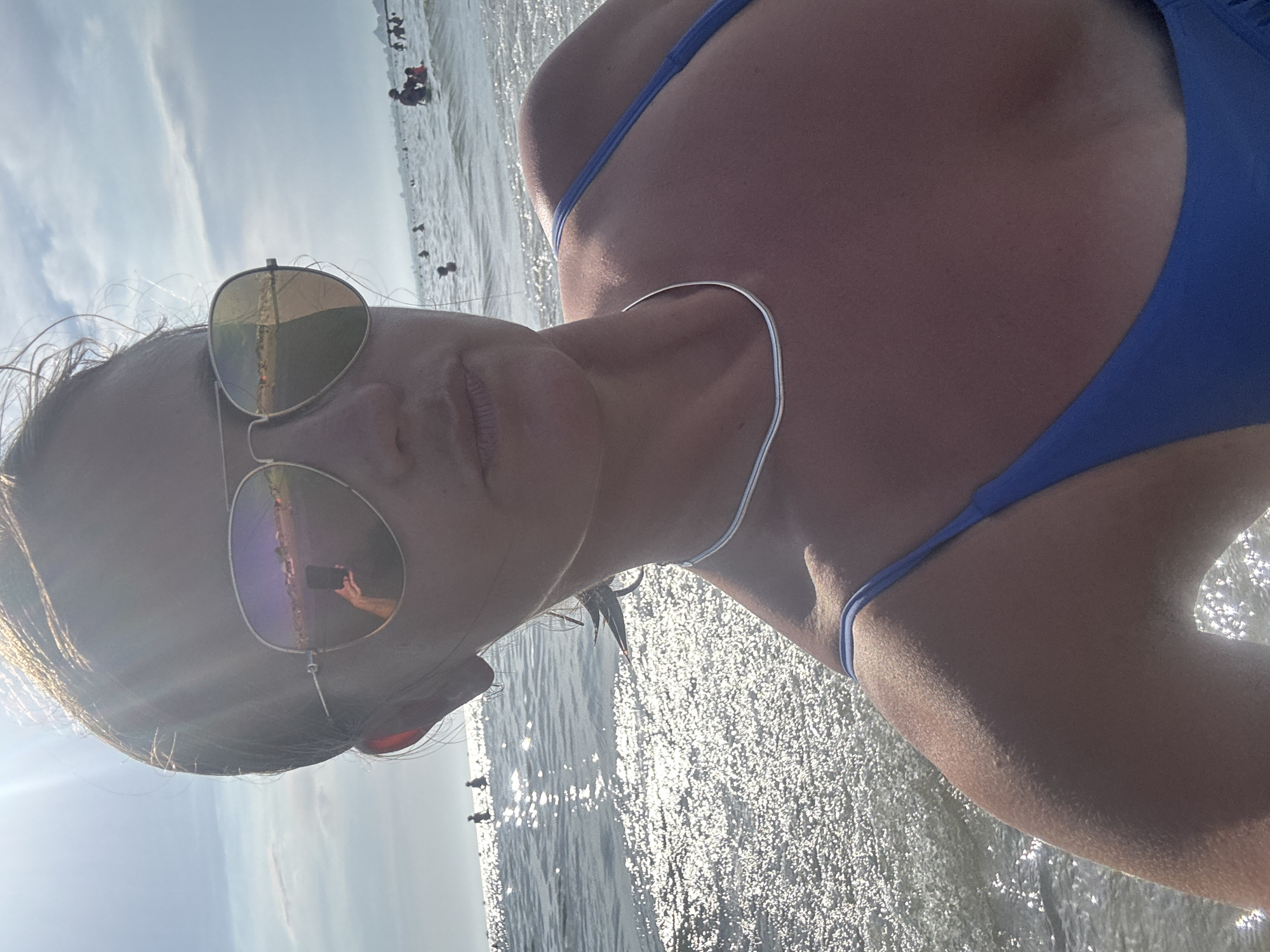 A woman is taking a selfie at the beach in Le Grau-du-Roi, France. She is wearing sunglasses, a silver necklace, and a blue bikini. The ocean is calm and there are people swimming and playing in the water. The sky is clear with some clouds and the sun is shining brightly. The reflection in the woman's sunglasses shows her holding a phone and taking the picture, with people and buildings visible in the background.