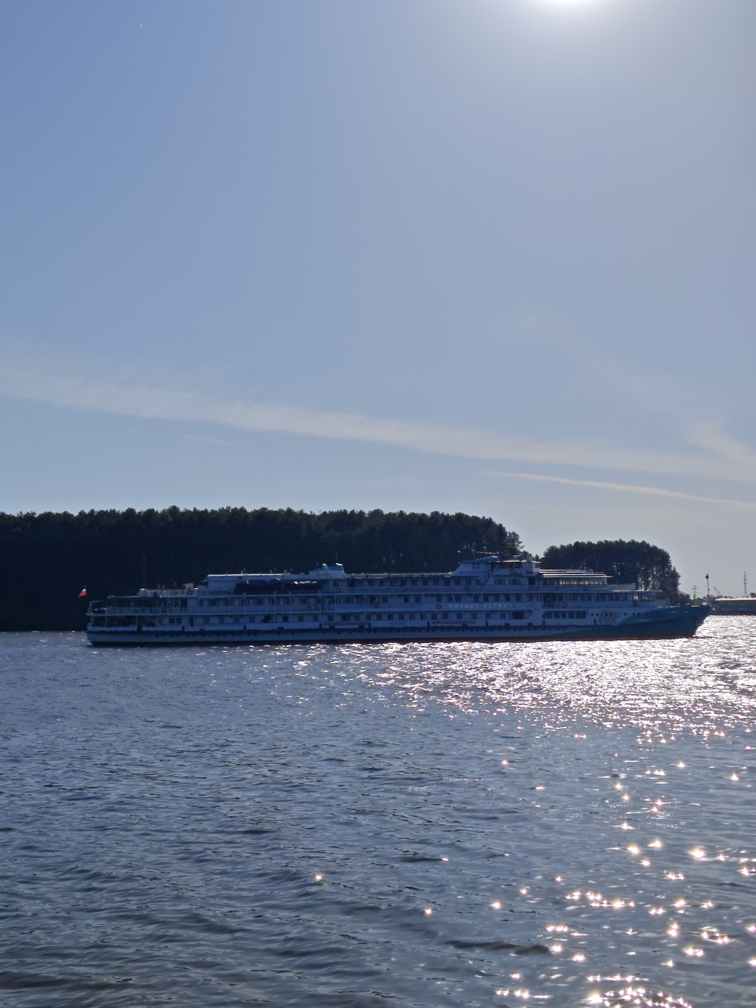 The image shows a multi-deck passenger cruise ship sailing on a body of water, possibly a river or lake, on a bright, sunny day. The ship is white with blue accents and features rows of windows. The surface of the water is reflective, shimmering with sunlight. In the background, there is a line of trees, likely a forest or park area. The clear blue sky occupies the majority of the upper portion of the image. The location is likely in the city of Chaikovsky, Russia.