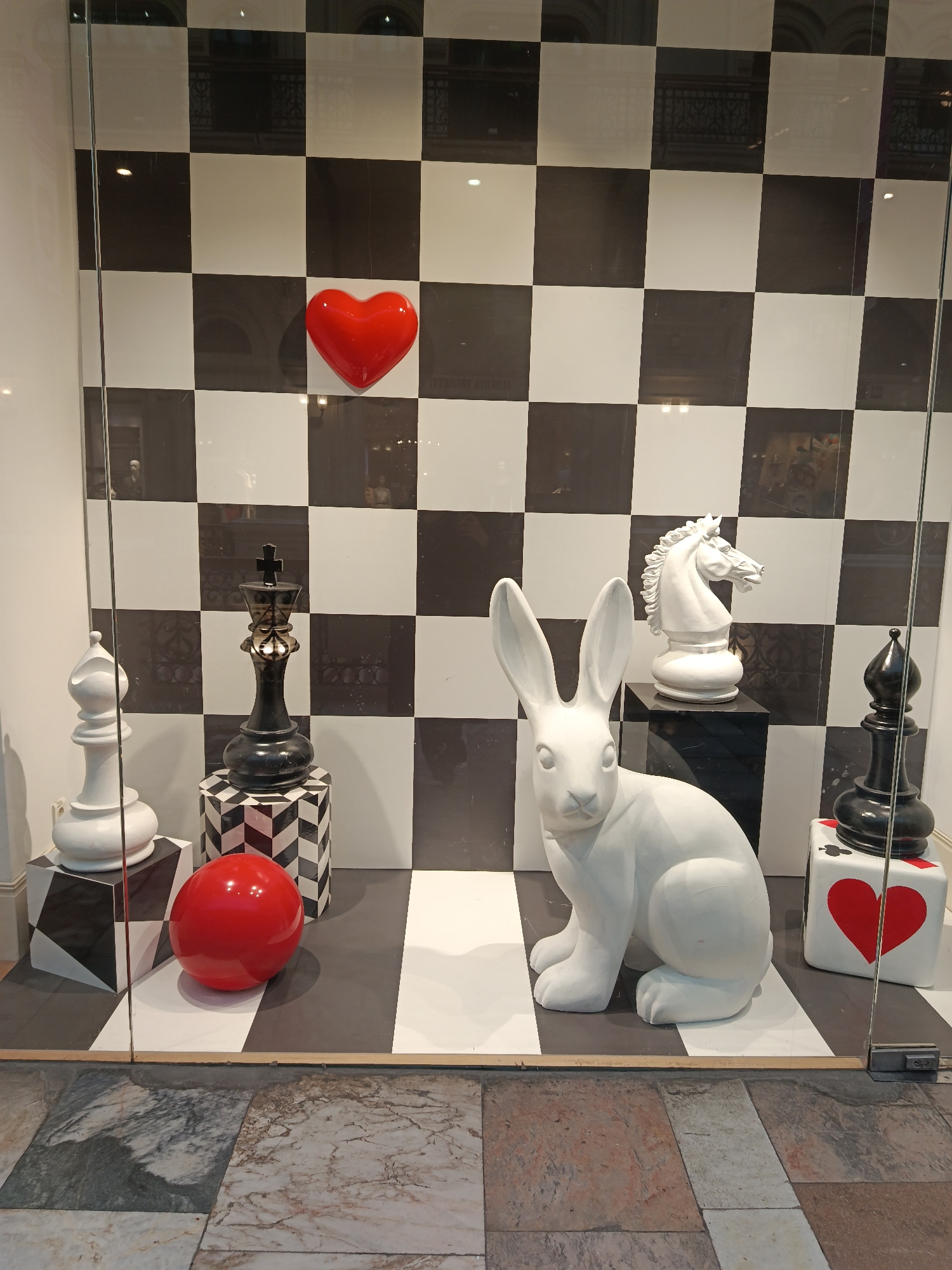 This media file captures a display window featuring an "Alice in Wonderland" or chess-themed art installation. The background and floor of the display are designed with a large black and white chessboard pattern.
The main subjects are several oversized sculptures. A large white rabbit sculpture, reminiscent of the White Rabbit from "Alice in Wonderland," sits prominently in the foreground on the right side. Surrounding it are giant chess pieces: a black king stands centrally on a black and white chevron-patterned pedestal, a white bishop is on the far left, a white knight on a black pedestal is behind the rabbit, and a black bishop is on the far right. The black bishop rests on a white cube pedestal adorned with a large red heart and a partial spade playing card symbol.
Additional objects include a bright red heart sculpture mounted on the checkered wall in the upper-middle section of the display, and a shiny red spherical ball resting on the checkered floor in the lower-left.
The display is viewed through a glass pane, which shows reflections of the exterior environment, including architectural details and lighting. The ground outside the display consists of large, multi-toned marble tiles. The color palette is primarily black, white, and red, creating a striking and whimsical aesthetic. No explicit text is clearly visible within the display.