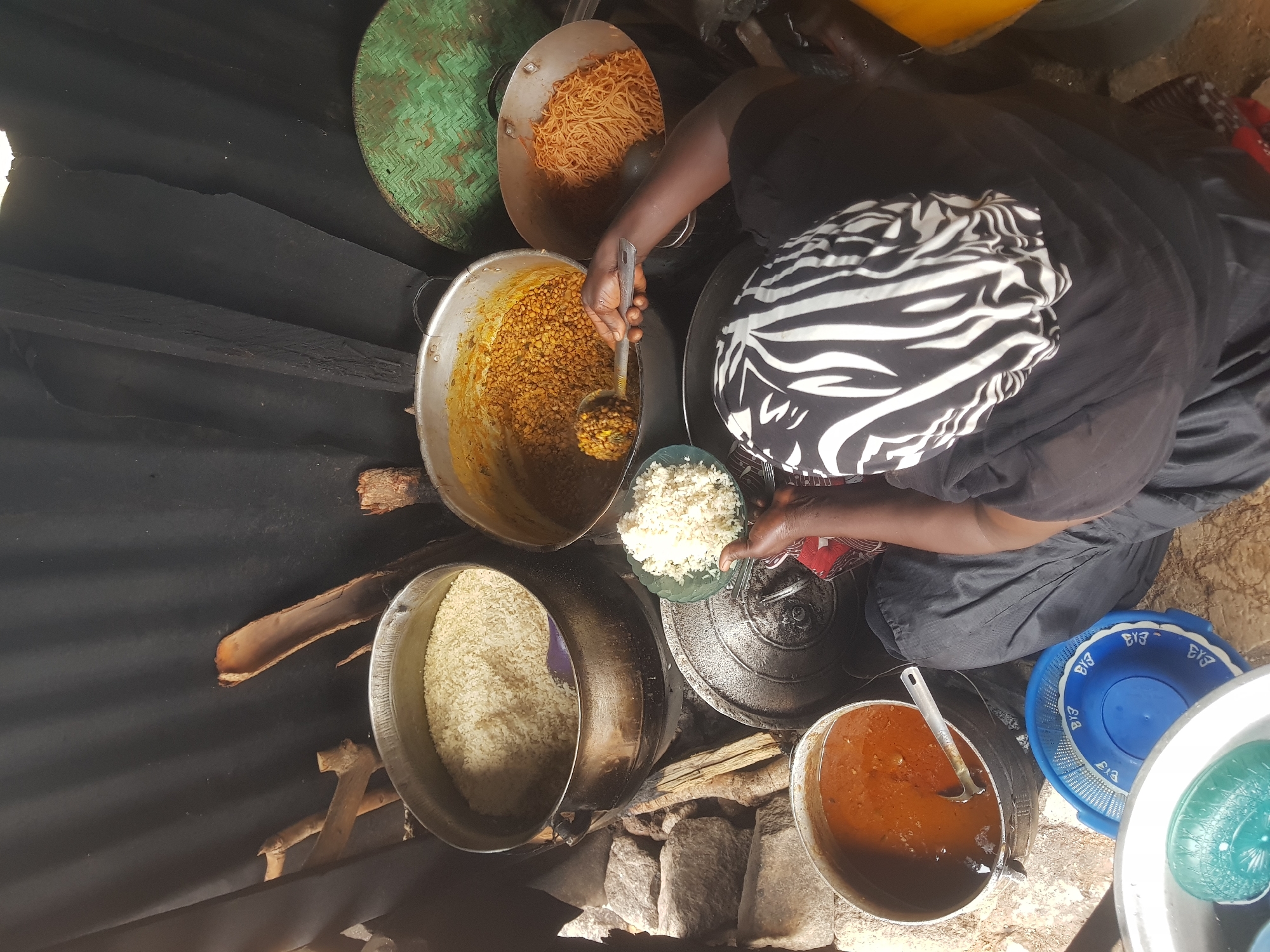 The overhead shot shows a busy food preparation scene, likely at an outdoor market stall, in the city of Jos, Nigeria. A person, wearing a black headscarf with a white zebra-like pattern and dark clothing, is crouched down and actively serving food. They hold a large ladle, scooping what appears to be a lentil or bean stew from a large pot into a bowl of white rice.
Surrounding the central action are several other large cooking pots. One pot is filled with a large quantity of cooked white rice. Another contains a dense, yellowish stew, possibly a groundnut stew or a similar local dish. A fourth pot holds a mound of cooked spaghetti or noodles. A smaller pot filled with a reddish-brown stew, likely a pepper soup or tomato-based sauce, is visible to the left, with a ladle resting inside.
The ground is a mix of rough stones and dirt. In the foreground, a blue plastic basket with white trim and a large silver bowl, which may contain more food or ingredients, are present. A green woven mat is visible in the background, along with some rough wooden branches and a dark, textured backdrop that could be a tarp or corrugated metal sheeting, suggesting a simple, open-air setup typical of many Nigerian markets. The overall impression is one of abundance and active food service.