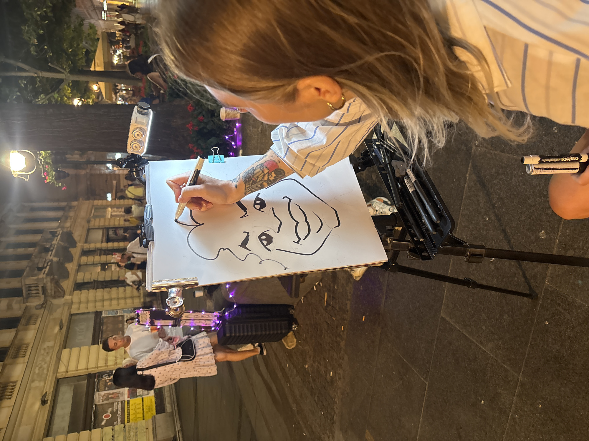 The image depicts an outdoor scene at night, likely in a pedestrian zone in Belgrade, Serbia, given the architectural style of the buildings and the overall ambiance. A caricature artist is in the foreground, from the shoulders down, drawing on a canvas placed on an easel. The artist has blonde hair and is wearing a striped shirt. They are holding a marker and appear to be in the process of drawing a cartoonish face.
Behind the artist, a couple is standing. The woman on the left is wearing a light-colored dress and carrying a black purse. The man next to her is wearing a white t-shirt. They are looking at the artist and the drawing. There is a black suitcase and some purple decorative lights near them.
The background shows a street with shops or restaurants, illuminated by streetlights and indoor lighting, creating a warm, inviting atmosphere. People are visible in the distance, suggesting a lively urban environment. A large tree is to the right of the easel, and a portable projector or light is attached to it, likely to illuminate the drawing area. The ground is paved with stone tiles. The overall impression is of a vibrant evening in a European city.