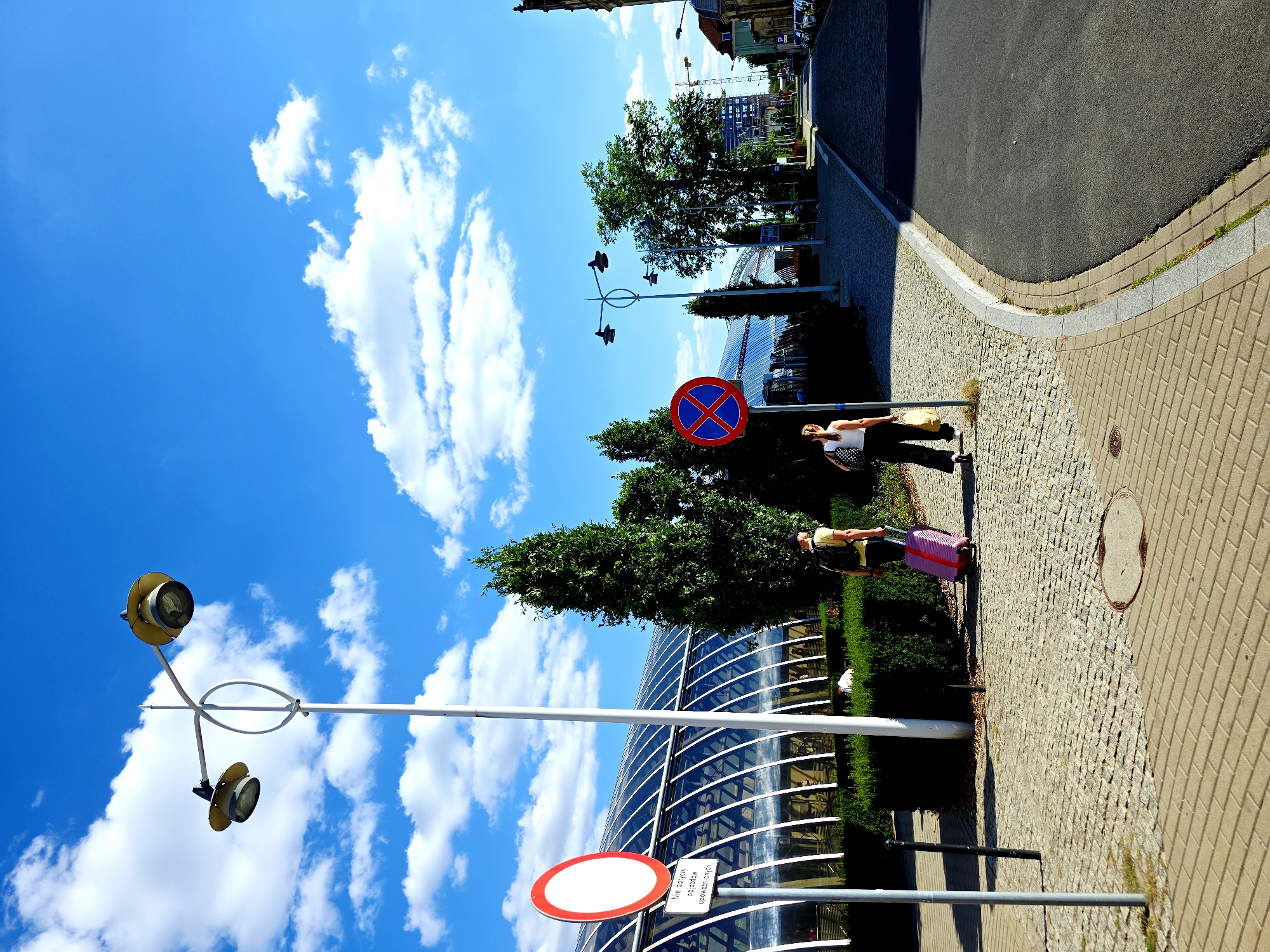 The image depicts a bright, sunny day with a blue sky and scattered white clouds. In the foreground, a paved sidewalk curves to the right, meeting an asphalt road. To the left of the sidewalk is a building with a distinctive curved, glass roof supported by white beams, possibly a modern architectural structure like a station or exhibition hall. Several streetlights with multiple lamps are visible along the sidewalk.
Two individuals are present. On the left, a person wearing a yellow t-shirt and dark pants, with a backpack, is seen from the back, pulling a bright pink suitcase on wheels. They are walking away from the viewer towards the right. Further to the right, another person, seen from the back, is wearing a white top and dark pants, carrying a shopping bag. They are also walking to the right.
Several traffic signs are visible. A red and white circular sign with a white horizontal bar is on the left, accompanied by a sign with Polish text. A red and blue circular sign with a white cross is further down the sidewalk to the right.
The environment includes manicured green hedges and a tall, slender evergreen tree. In the distance, more buildings and trees line the street, and there are cars parked along the right side of the road. The overall scene suggests a public area in a city, likely a transit hub or a commercial district. The specific location is the vicinity of the Poznań Główny train station in Poznań, Poland.