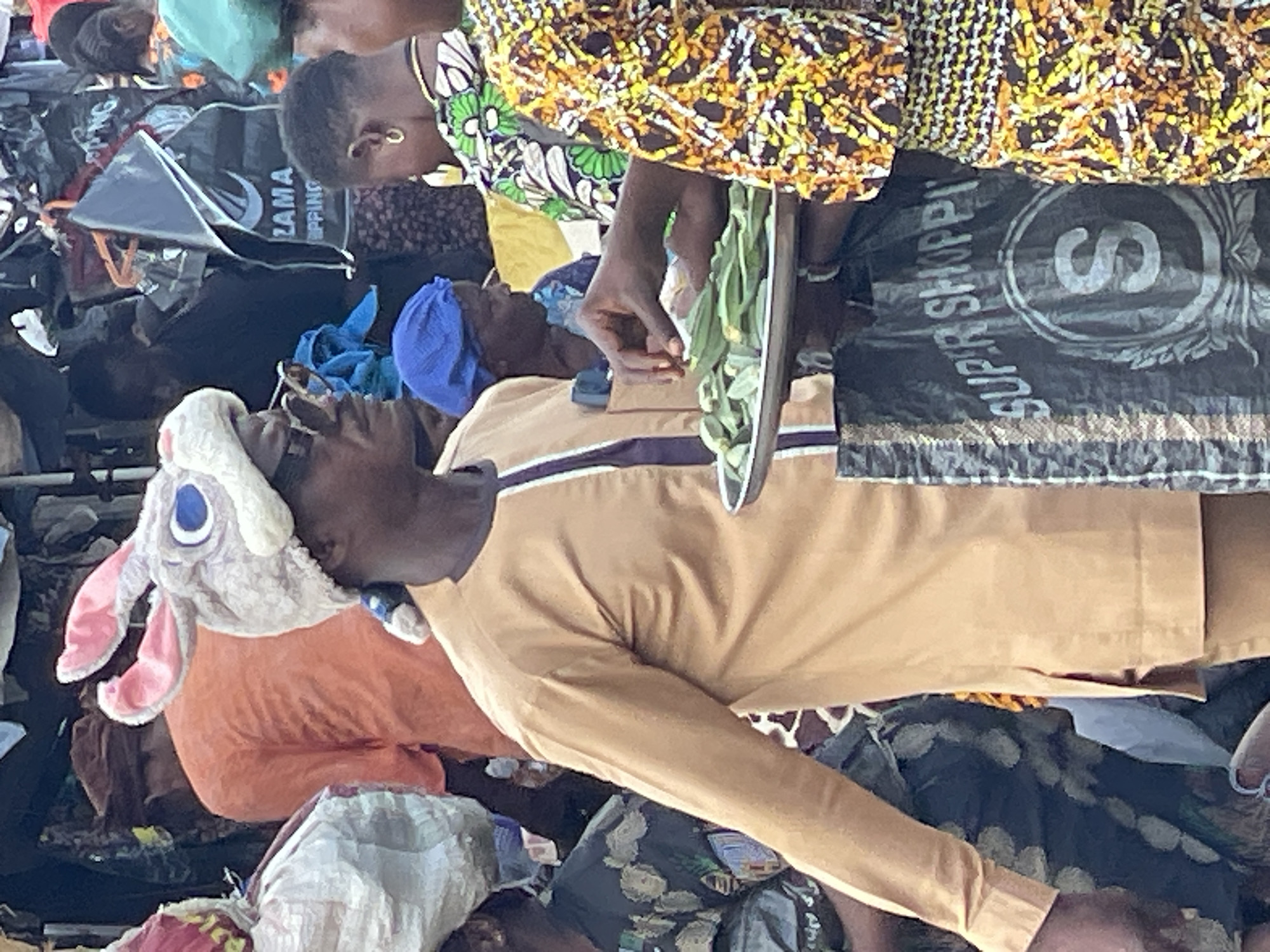 This image captures a bustling open-air market scene in the city of Unknown, Unknown. The central figure is an adult male, seen in profile, facing left. He has dark skin, wears dark sunglasses, and an unusual white and pink bunny ear hat featuring large blue cartoon eyes and a pink nose. A dark blue fabric is tied at the back of his neck, possibly integrated with the hat. He is dressed in a light brown (tan/ochre) long-sleeved tunic with a dark purple trim along the placket and collar. He holds a black "SUPER SHOPPING" branded bag with a large white "S" logo.
To his right, a woman with dark skin, wearing a brightly patterned dress (yellow, orange, black, white abstract/geometric pattern), extends her right hand over a silver-colored metal tray filled with green okra pods. A gold hoop earring is visible in her right ear. Behind her, a partially visible person wears a blue headwrap. Further in the background, another person, with their back to the viewer, wears an orange-colored shirt. Various other indistinct figures populate the crowded background. Hanging in the mid-ground, a dark grey/black bag is visible, displaying "ZAMA SHIPPING" in white text. The overall environment suggests an active commercial area with multiple individuals engaged in activities typical of a marketplace.