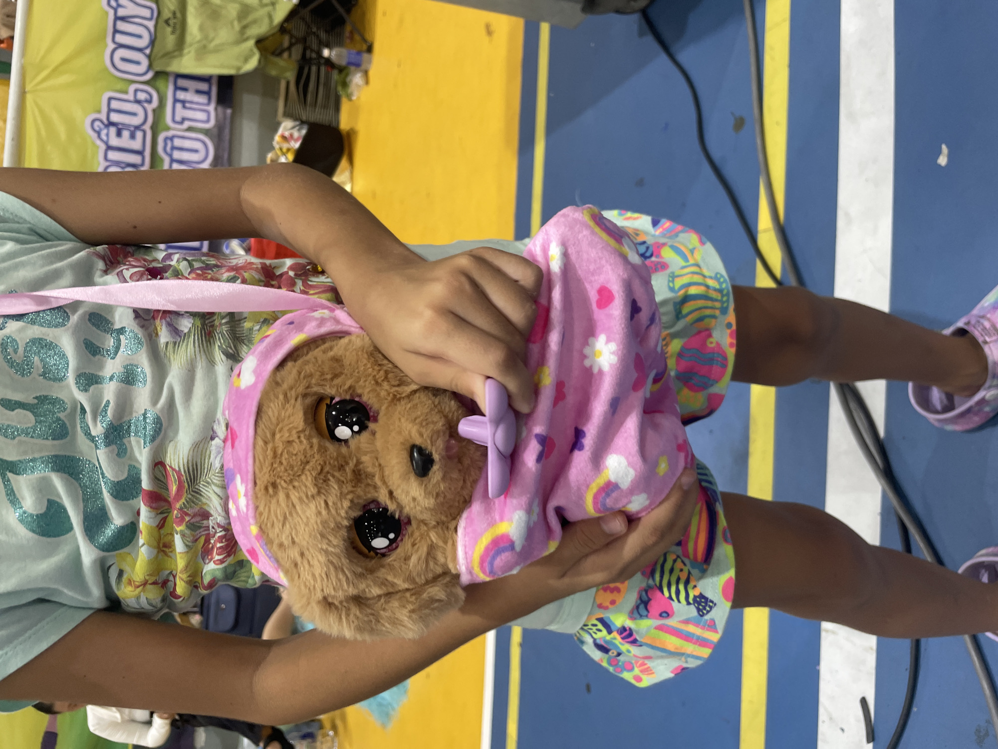 In Thuận An, Vietnam, a young girl is captured in a close-up shot, holding a plush toy dog. She is wearing a light blue t-shirt with a colorful floral print and a brightly patterned skirt. The plush dog, with large, expressive eyes and a brown furry body, is wrapped in a pink blanket adorned with rainbows, hearts, and flowers. The girl's hands are visible, gently holding the toy. The background suggests an indoor environment with a bright yellow floor marked with blue and white lines, possibly a gymnasium or recreational area. A partial view of a banner with Vietnamese text is also visible in the background. The overall scene conveys a sense of childhood playfulness and affection.