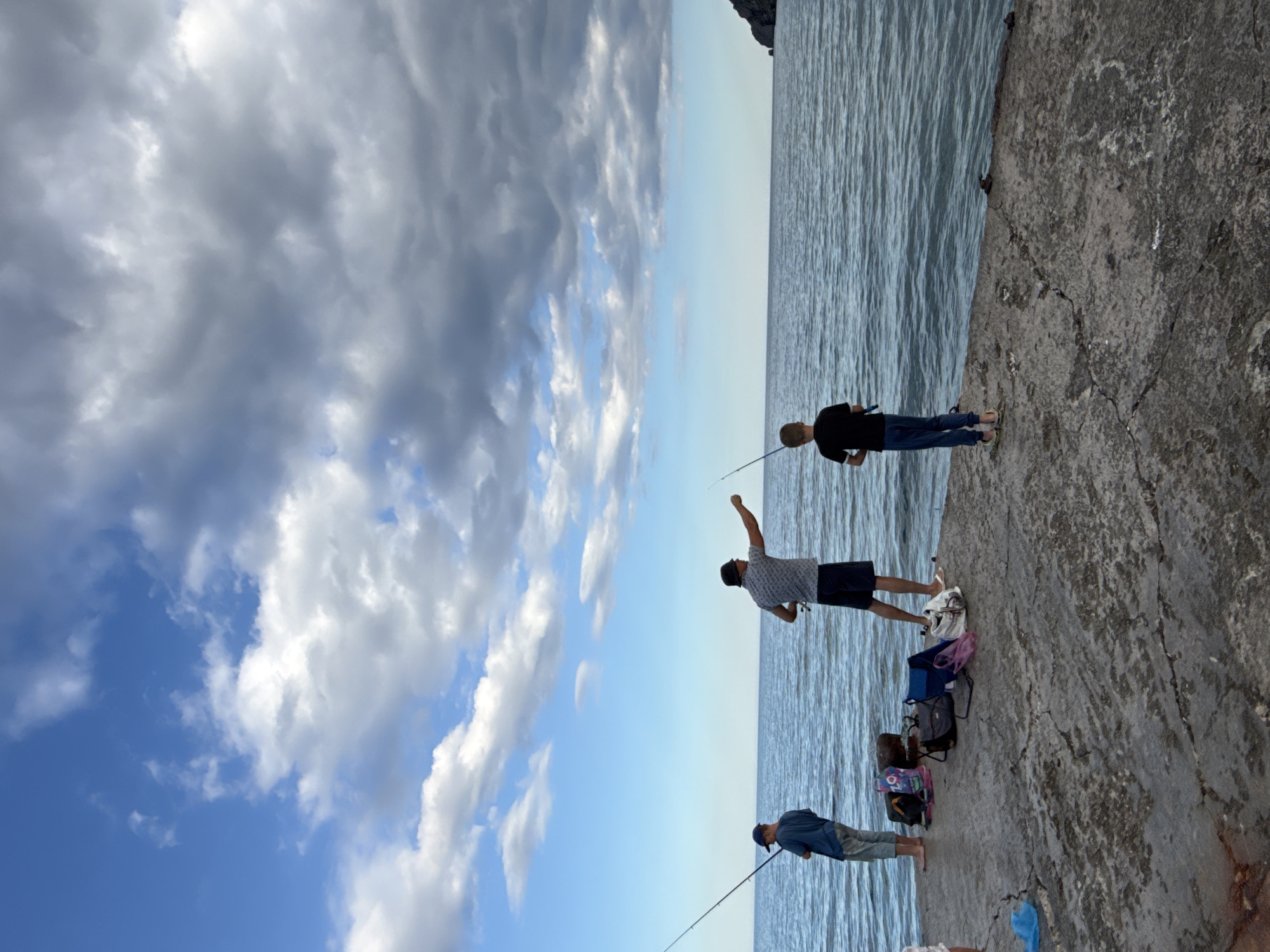 Three individuals, likely two adults and one child, are fishing from a concrete pier extending into a body of water, presumably the Black Sea. The sky is partly cloudy, with dramatic grey clouds dominating the upper portion of the frame. The sea is a deep blue with visible ripples, and a distant, dark, rocky landmass is visible on the right horizon. The pier is weathered and cracked, with several bags and fishing equipment placed on it. The individuals are positioned at intervals along the pier, each holding a fishing rod. One adult, wearing a dark hat and patterned shirt, is in the center, seemingly casting a line. Another individual, a child in a black t-shirt and blue pants, stands to the right, also fishing. To the left, a third person, a younger individual in a blue shirt and light-colored pants, is also fishing. The overall scene depicts a leisure activity in an outdoor, coastal environment.