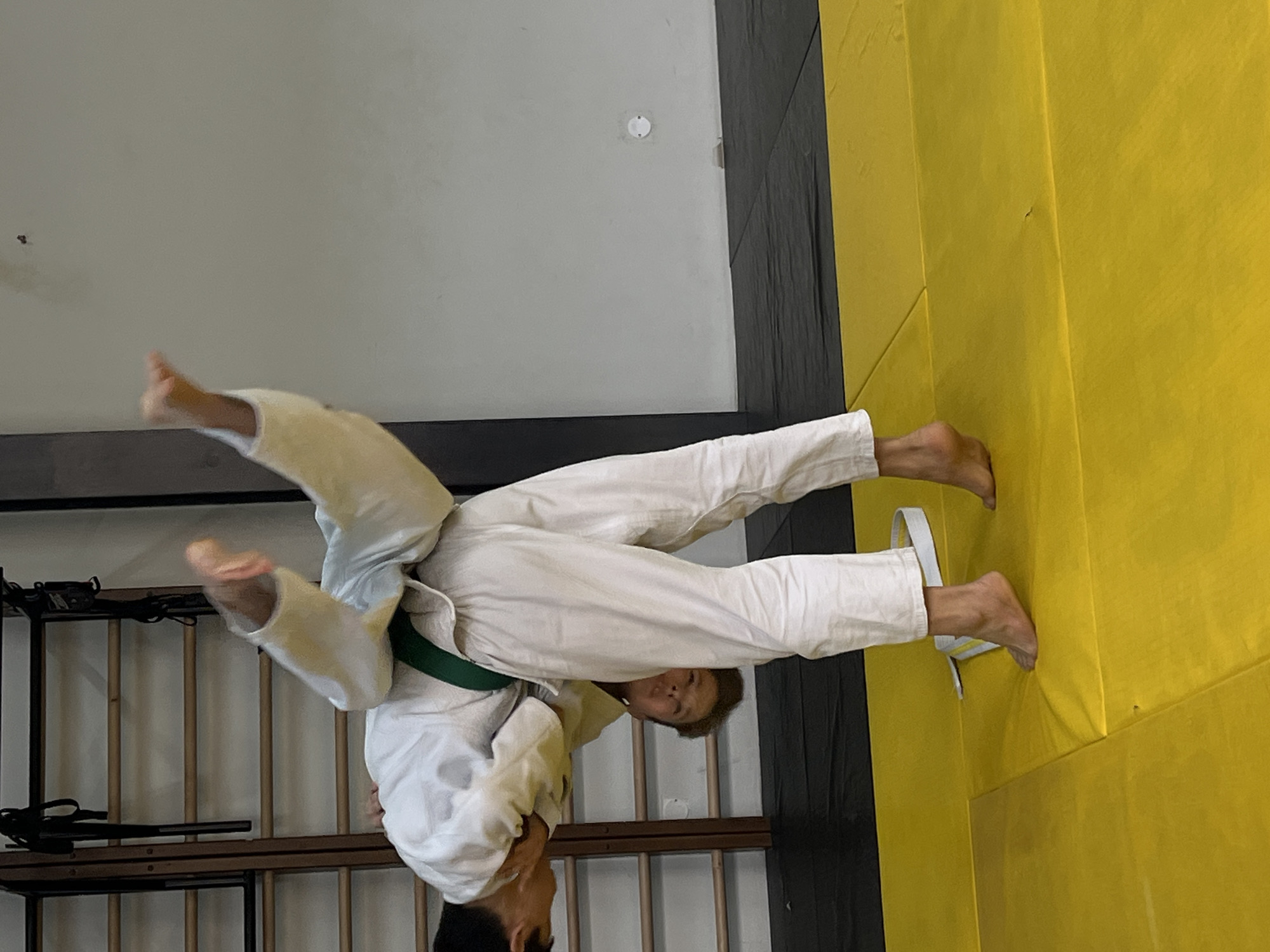 Two individuals are engaged in a judo training session. One individual, wearing a white gi with a green belt, is performing a throwing technique on another individual, whose face is obscured. The scene is set in a dojo with black and yellow mats. A wall is visible in the background, with a storage rack. There is no indication of the city of Krasnoyarsk.