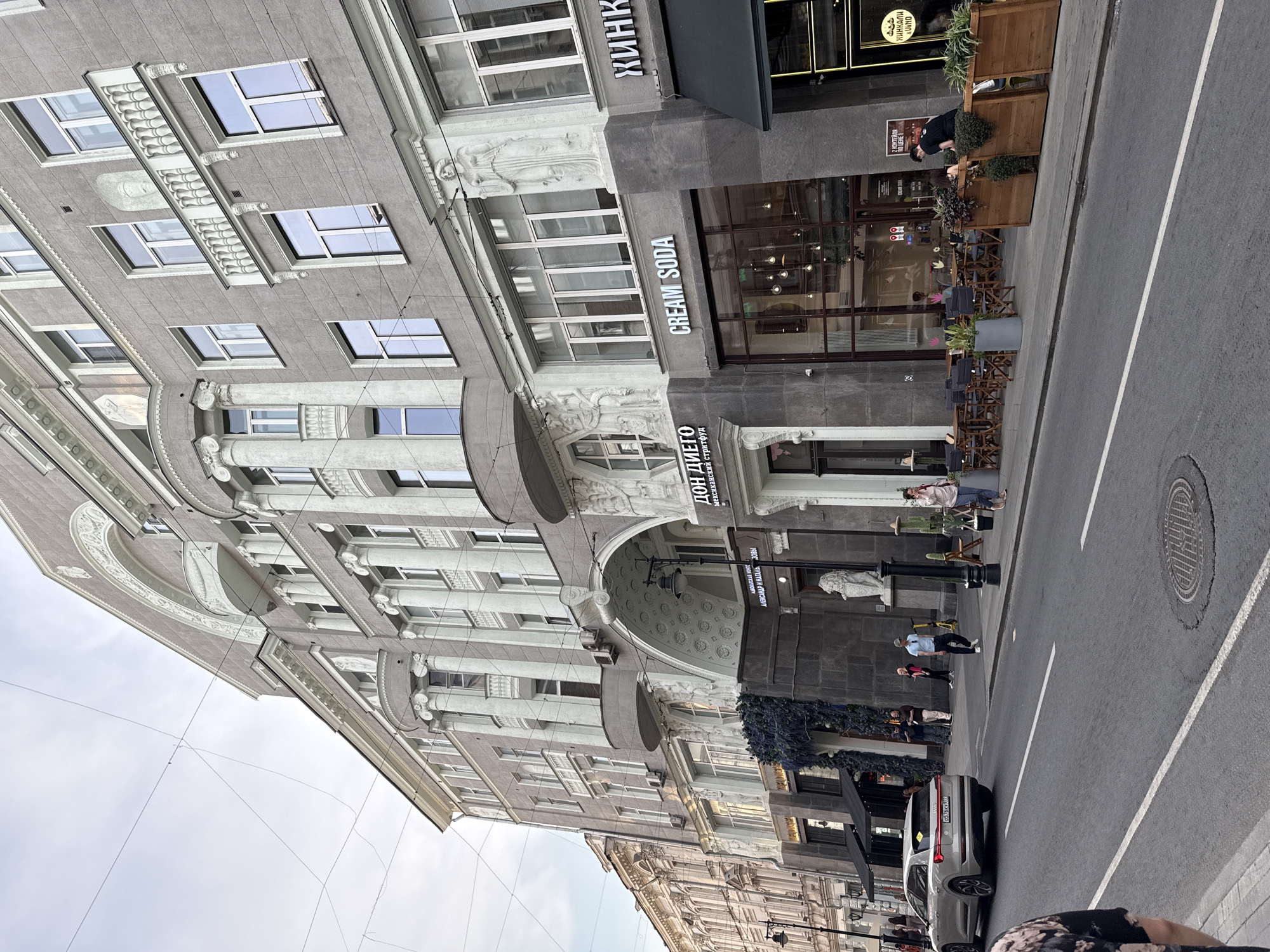 The image captures a street scene in Moscow, Russia, featuring a grand, ornate building with classical architectural elements. The building is light gray and adorned with columns, sculptures, and intricate detailing above the windows and along the roofline. The ground floor houses businesses, including "DON DIEGO" which appears to be a Mexican food establishment, and "CREAM SODA," a cafe or bar. An Audi e-tron electric car is parked on the left side of the street, which is paved and has white lane markings. Several pedestrians are visible on the sidewalk, some dining at outdoor tables set up in front of "CREAM SODA." The sky is overcast, suggesting a mild or cloudy day. The scene conveys a sense of urban life with a blend of historic architecture and modern commercial activity.