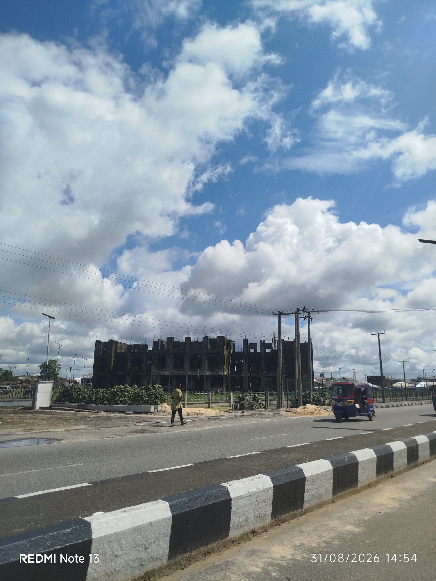 The image shows a construction site in Yenegoa, Nigeria, with a partially built structure dominating the center. The sky is filled with large, white cumulus clouds against a bright blue backdrop. A single person, wearing a yellow and brown patterned shirt and dark trousers, walks across the asphalt road in front of the construction. To the right of the road, a red and blue three-wheeled vehicle, commonly known as a "Keke Napep" or "Marwa", is visible. The road itself has a painted median with alternating black and white stripes. Power lines are strung across the upper portion of the image, supported by poles. The overall scene suggests a bright, sunny day in an urban or semi-urban area undergoing development.