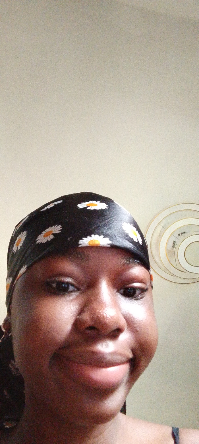 The scene is set indoors in Umudike, Nigeria. A close-up, frontal portrait captures an individual with dark skin, from the forehead to the upper chest. The person wears a black headscarf patterned with white daisies that have yellow centers. A subtle smile is present, and the subject looks directly into the camera. There is visible swelling on the right side of the face, specifically around the cheek and jaw area. A dark strap of clothing is partially visible on the lower right. The background consists of a plain, light-colored wall, with a circular, decorative wall hanging featuring concentric rings visible on the upper right.