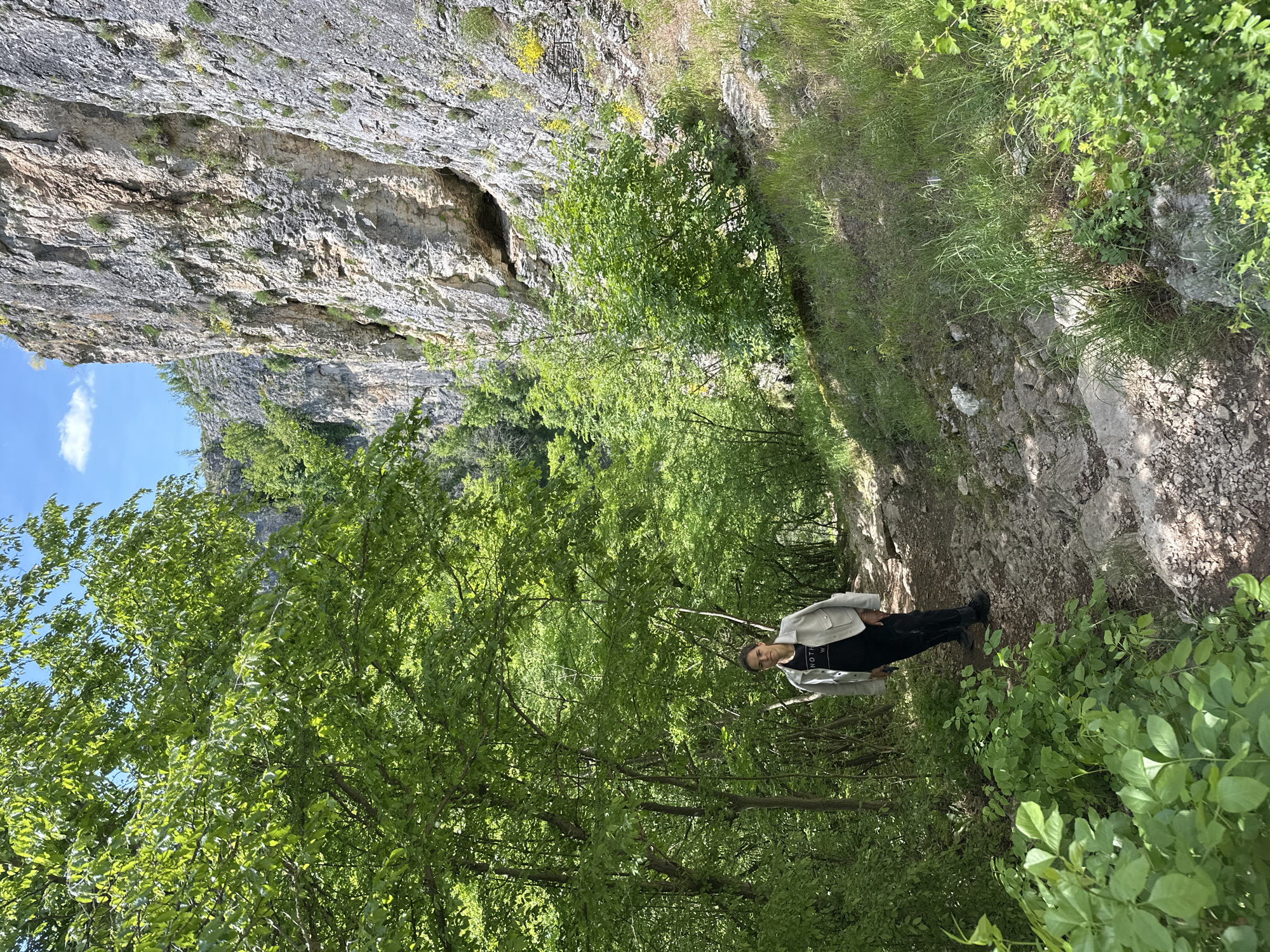 The image shows a person standing on a dirt path in a wooded area, with a rocky cliff face to their left. The person is wearing black pants, a black t-shirt, and a light-colored jacket. The path is surrounded by trees and foliage. The cliff face is gray and rocky, with some vegetation growing on it. The sky is visible in the upper left corner of the image and appears to be a clear blue. The photo appears to be taken in Karlukovo, Bulgaria, as that was the specified location.