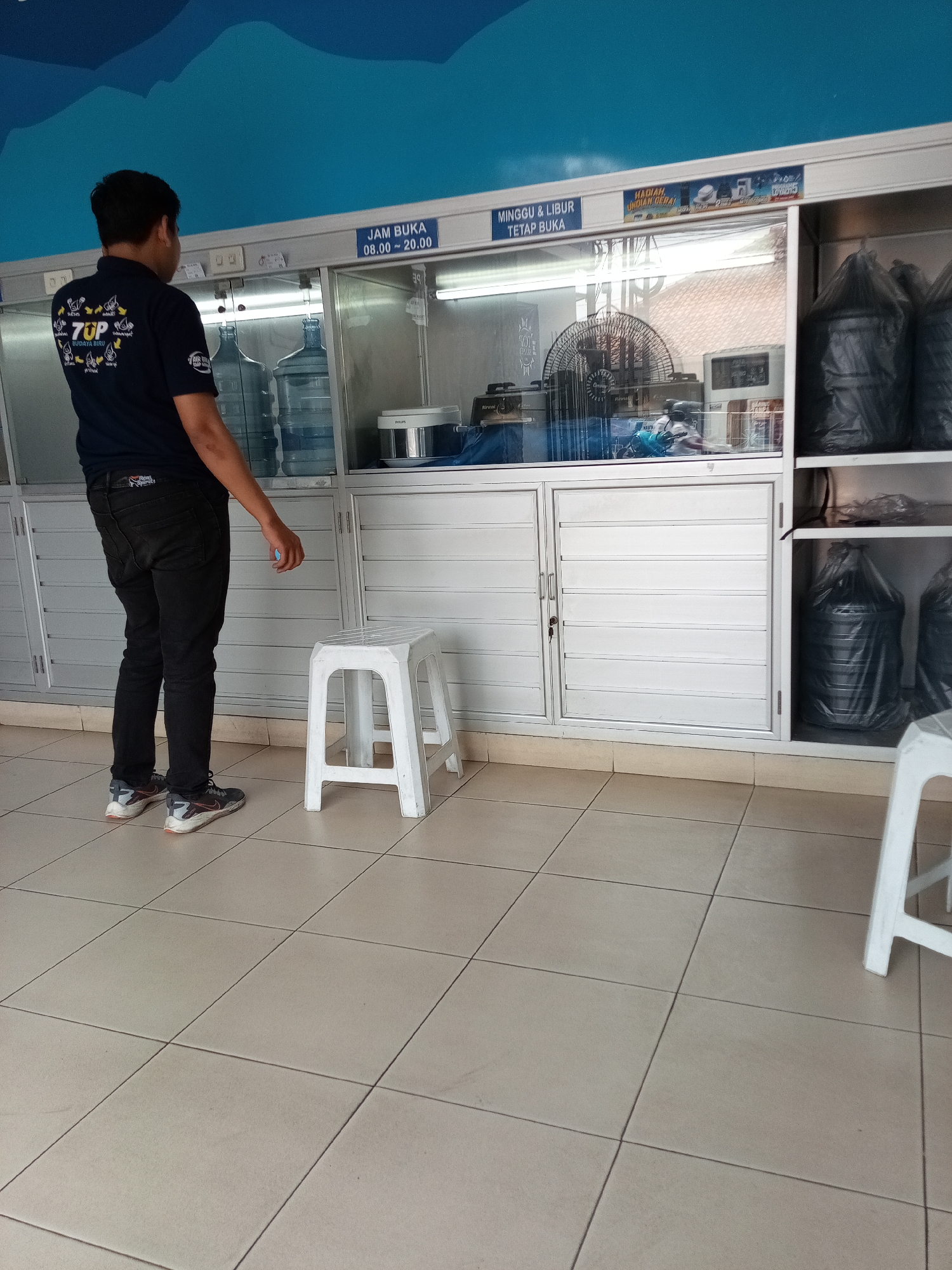 The image depicts an indoor scene at what appears to be a water refill station in Cimahi, Indonesia. A person, seen from behind and to the left, is wearing a dark blue t-shirt with a "7UP" logo and black jeans. They are standing on a tiled floor and looking towards a counter area.
The counter is behind a clear glass partition and is lined with several large water dispenser bottles, some full and some appearing empty. Above the counter, there are signs indicating operating hours. One sign reads "JAM BUKA 08.00 - 20.00", meaning "OPENING HOURS 08:00 - 20:00". Another sign, partially visible, states "MINGGU & LIBUR TETAP BUKA", which translates to "SUNDAY & HOLIDAYS STILL OPEN". There are also promotional materials visible above the counter.
Behind the partition, equipment such as a fan and other electrical appliances are present. To the right of the counter are shelves stocked with large black trash bags, some containing what appear to be water containers. A white plastic stool is positioned on the tiled floor to the right of the person. The background features a wall with a blue wavy pattern at the top. The overall environment suggests a commercial establishment focused on water dispensing services.